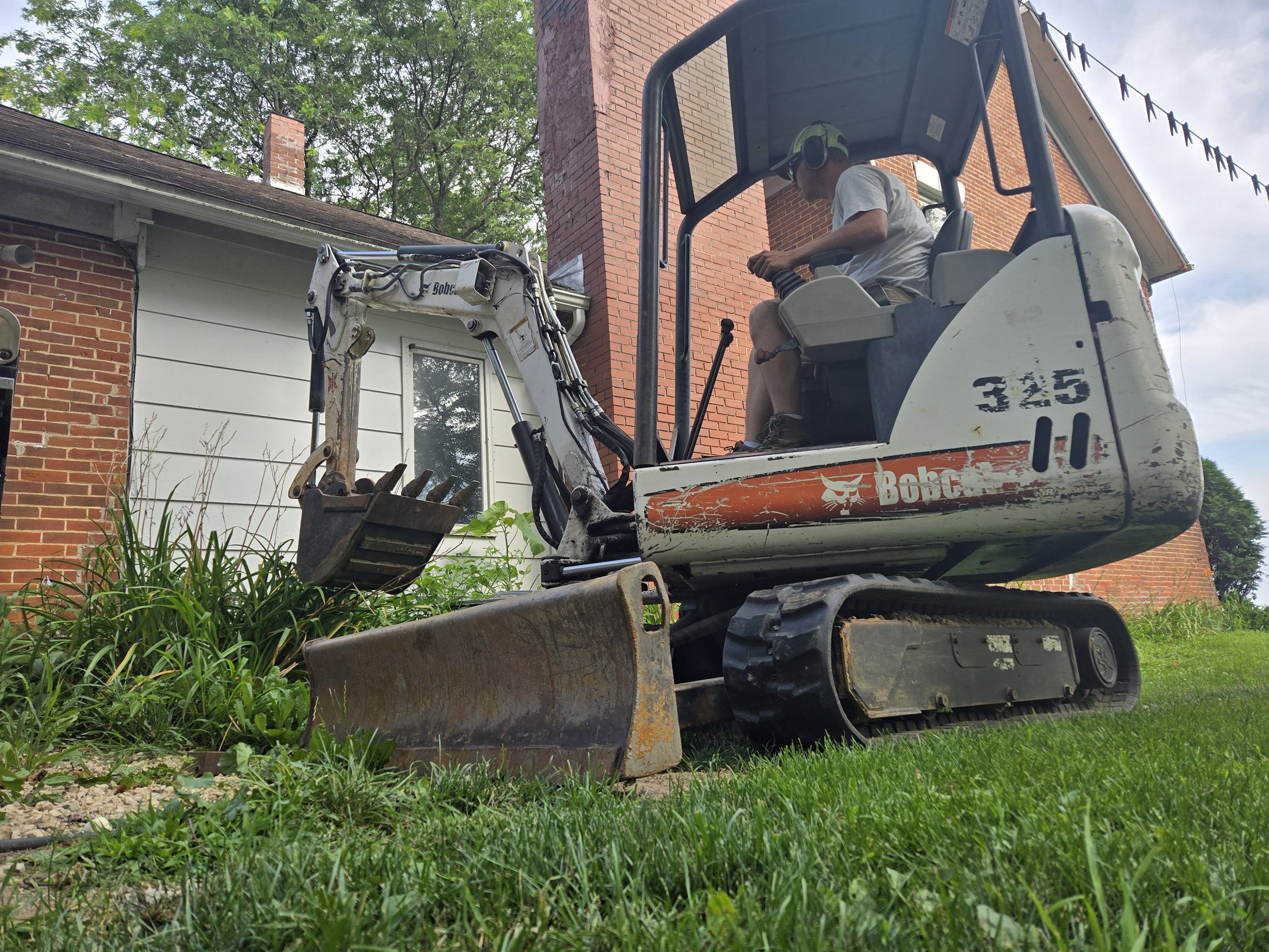 A man operating a small Bobcat excavator in a backyard, clearing away plants near a house with white and brick walls.