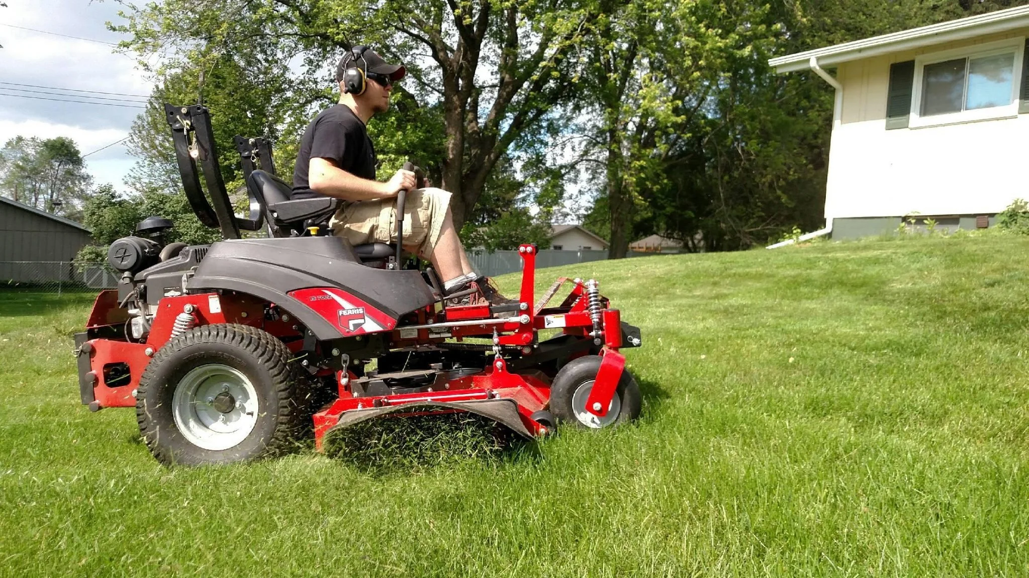 A person operating a red riding lawn mower on a grassy lawn. The person is wearing a black shirt, beige cargo shorts, a cap, and ear protection. There are trees and a house in the background.