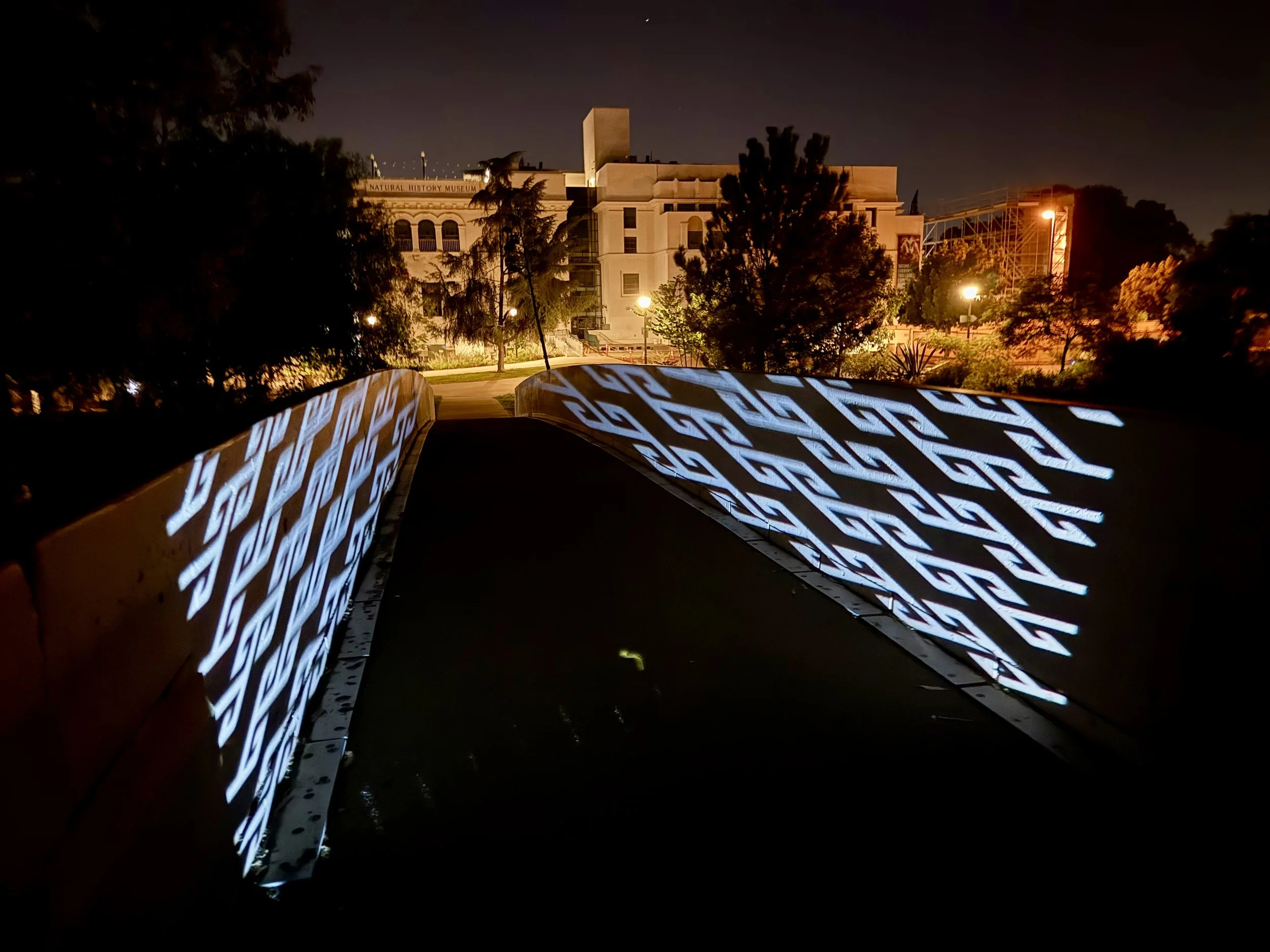 Nighttime view of a walkway in San Diego Balboa Park with illuminated patterned railings, trees, and a building in the background.