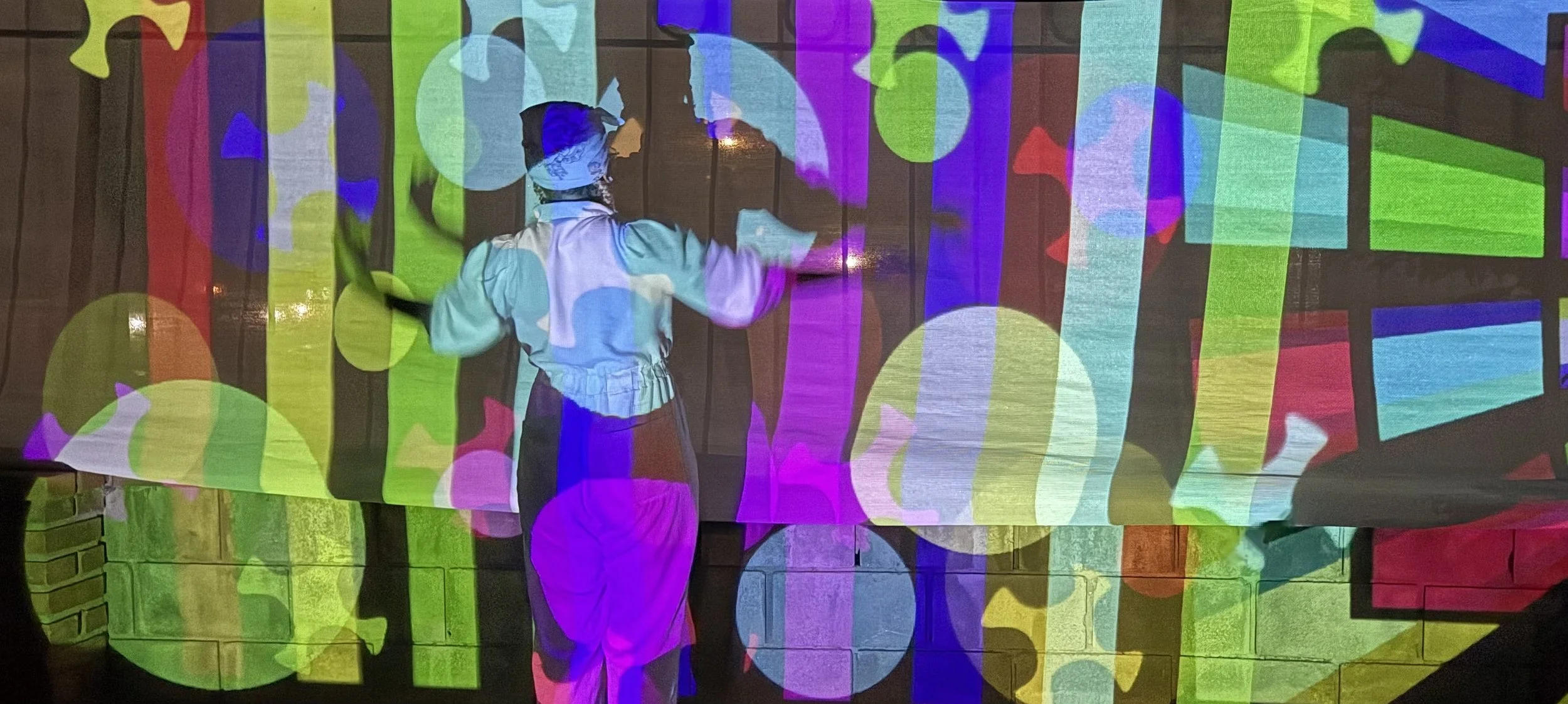 A person standing in front of a brick wall with colorful digitally projected circles and geometric shapes of various sizes and bright colors, creating an abstract visual effect.