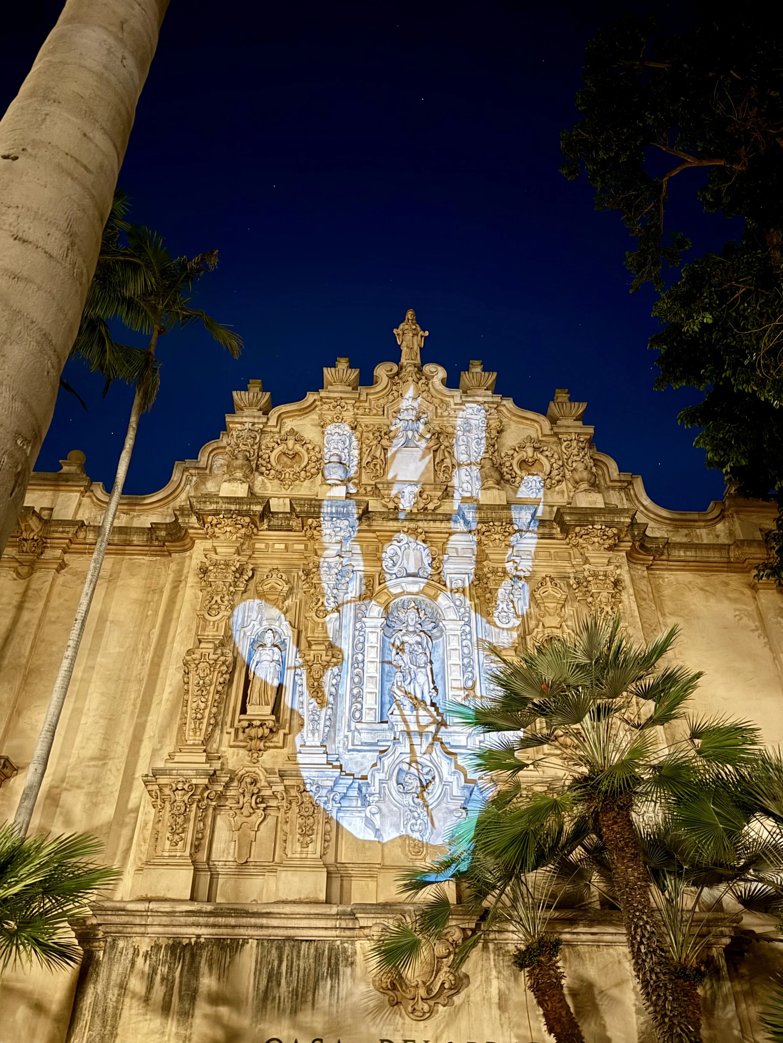 Night view of a decorated building facade with light projections in San Diego Balboa Park, featuring stone statues and ornate architectural details, surrounded by trees and palm trees against a dark sky.