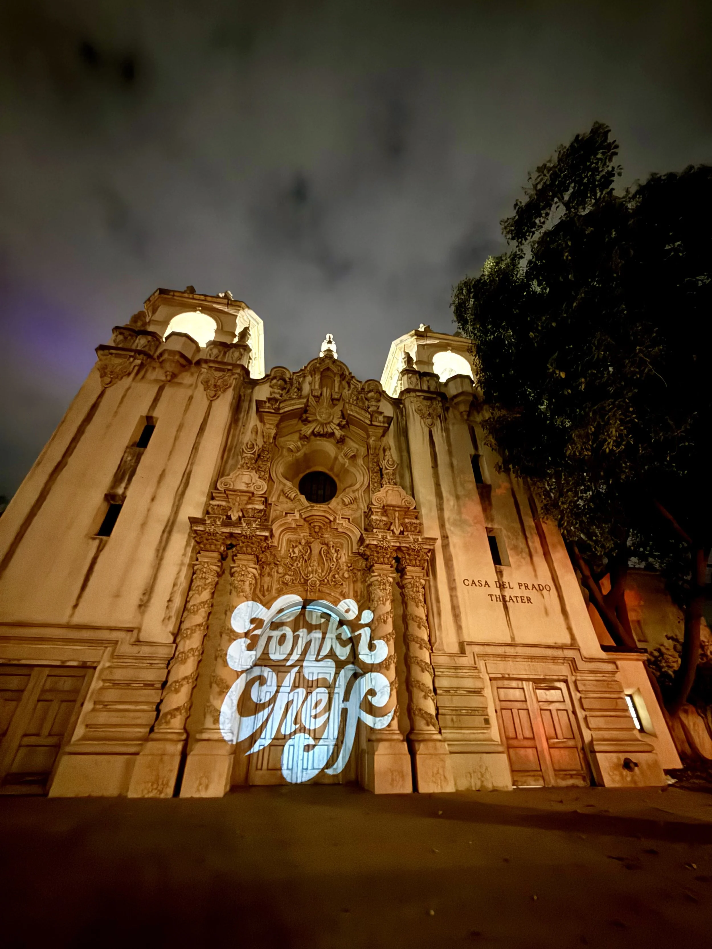Nighttime view of Casa del Prado Theater in San Diego Balboa Park with graffiti projection reading 'Fonki Cheff' on its ornate facade.