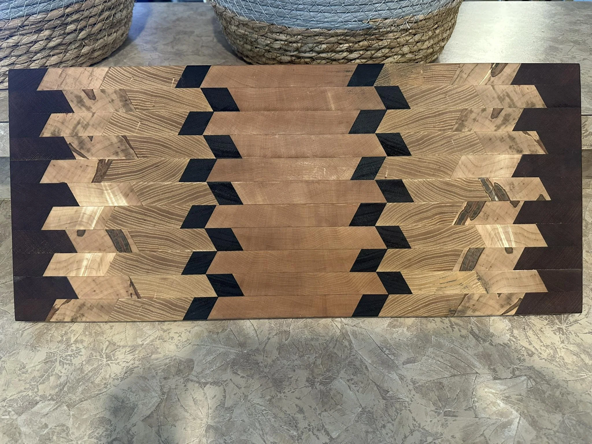 Close-up of a geometric wooden table with alternating light and dark wood patterns, with woven baskets in the background.