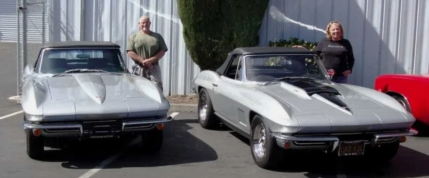 Two vintage Corvette cars parked side by side with two people standing behind each car, posing for the photo.