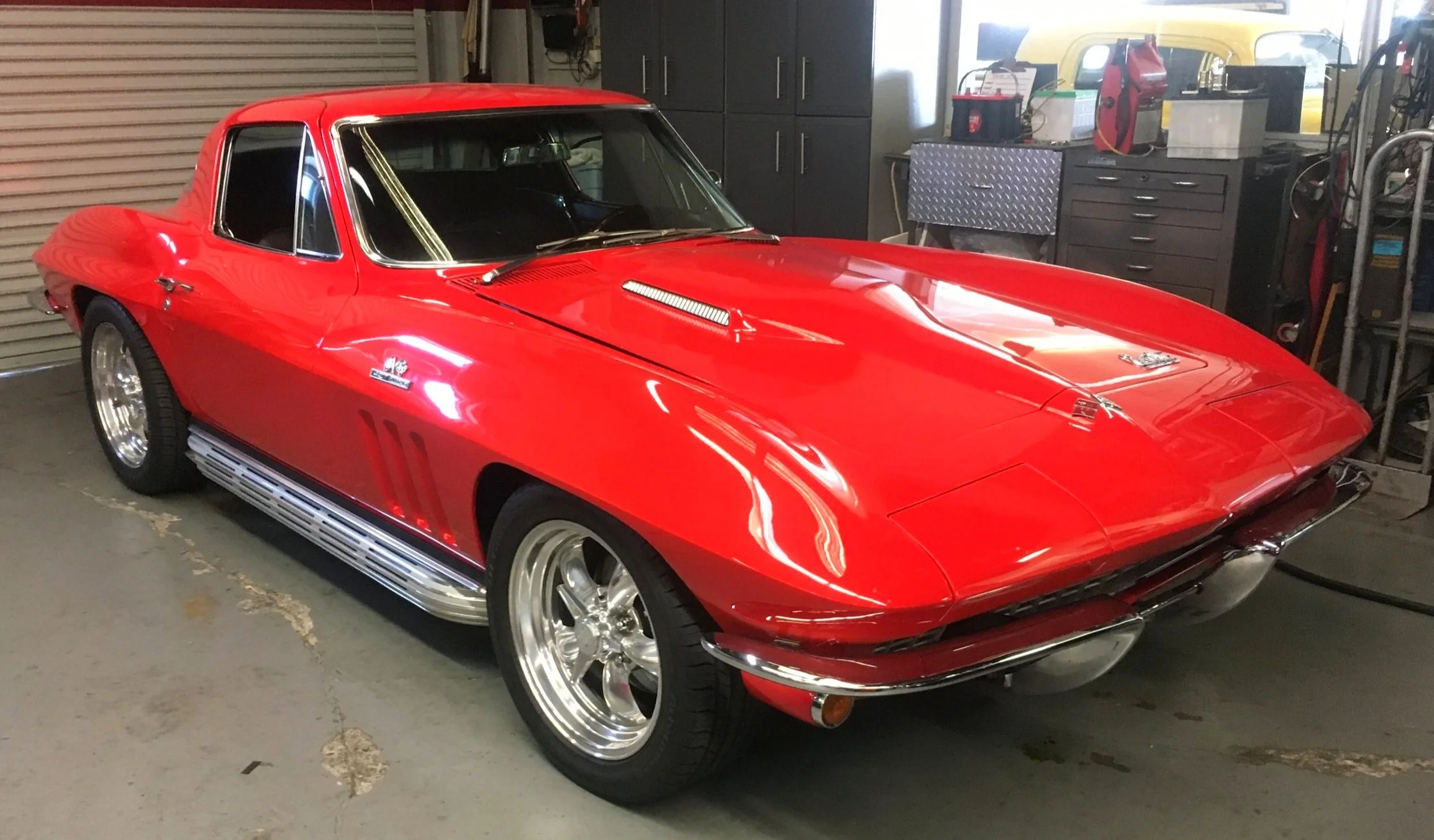 Red classic Chevrolet Corvette parked in a garage with tools and equipment in the background.