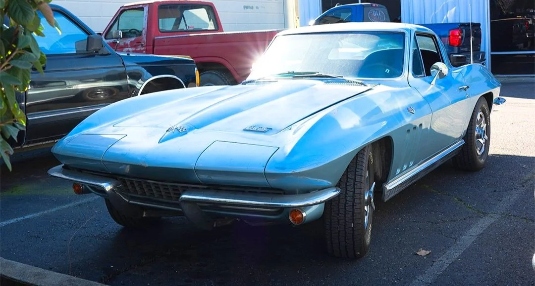 A light blue vintage Chevrolet Corvette parked in a lot next to other vehicles, with reflections of the surrounding area on its shiny surface.