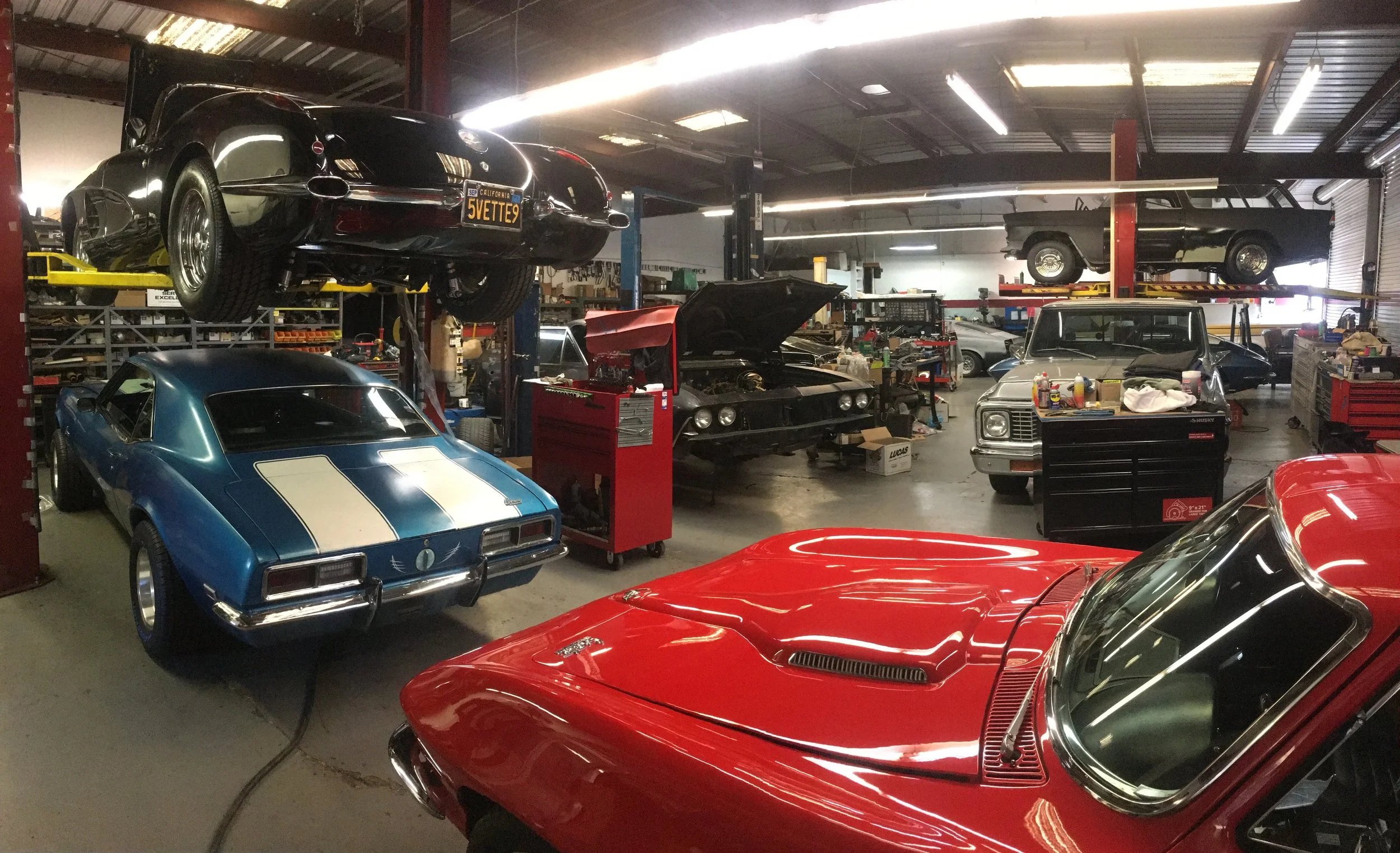 Interior of a car workshop with multiple classic cars, some on lifts and others parked or being worked on, including a black sports car on a lift, a blue muscle car, a red car, and an older pickup truck.
