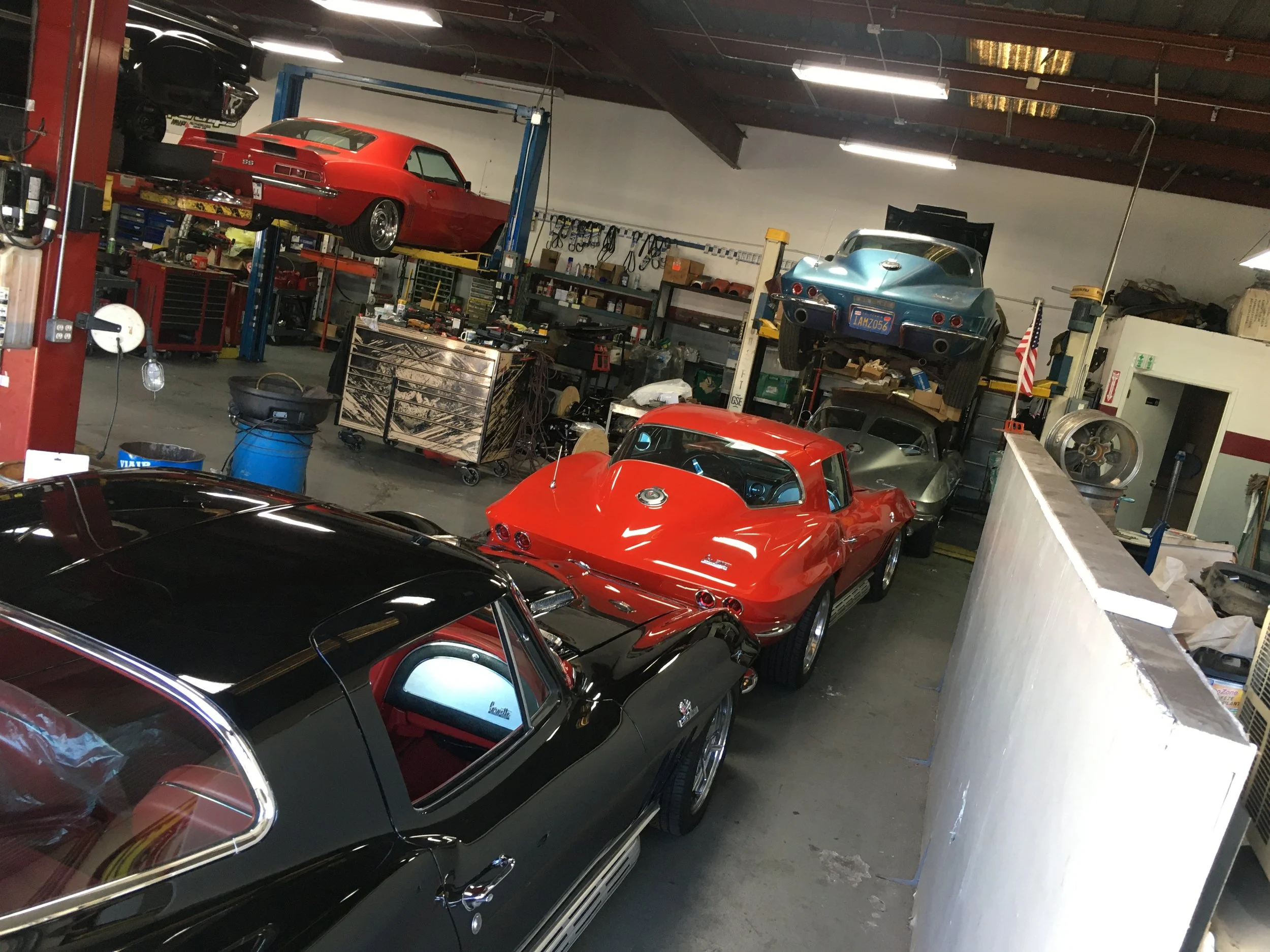 A garage filled with various classic and vintage cars, including a black car in the foreground, a red car behind it, and several others mounted on lifts and parked inside the workshop.