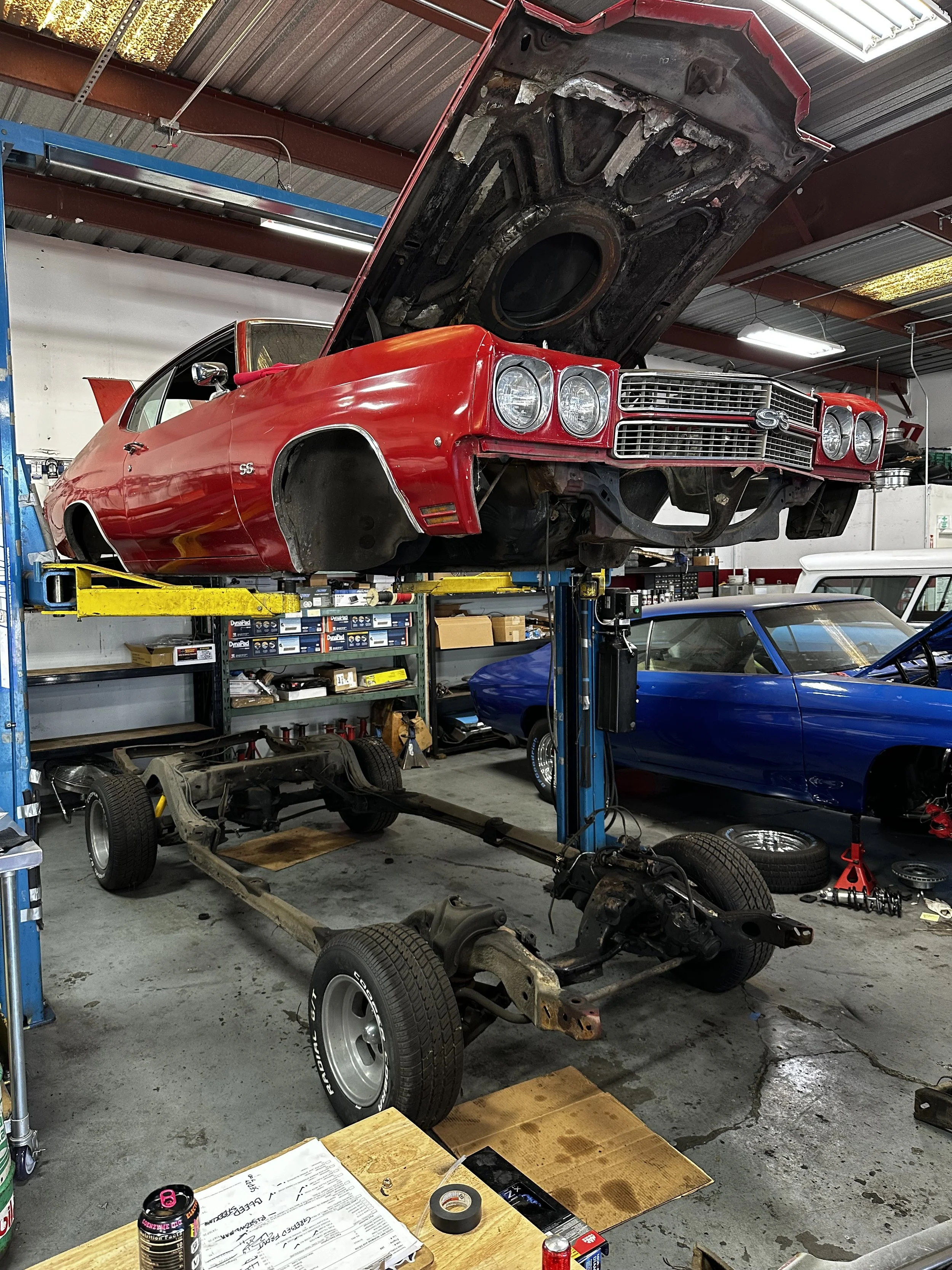 A red vintage Chevrolet Chevelle SS car elevated on a lift in a garage, partially disassembled with wheels removed, and an engine bay open with the hood raised. There are two other classic cars parked nearby, one blue and one white.