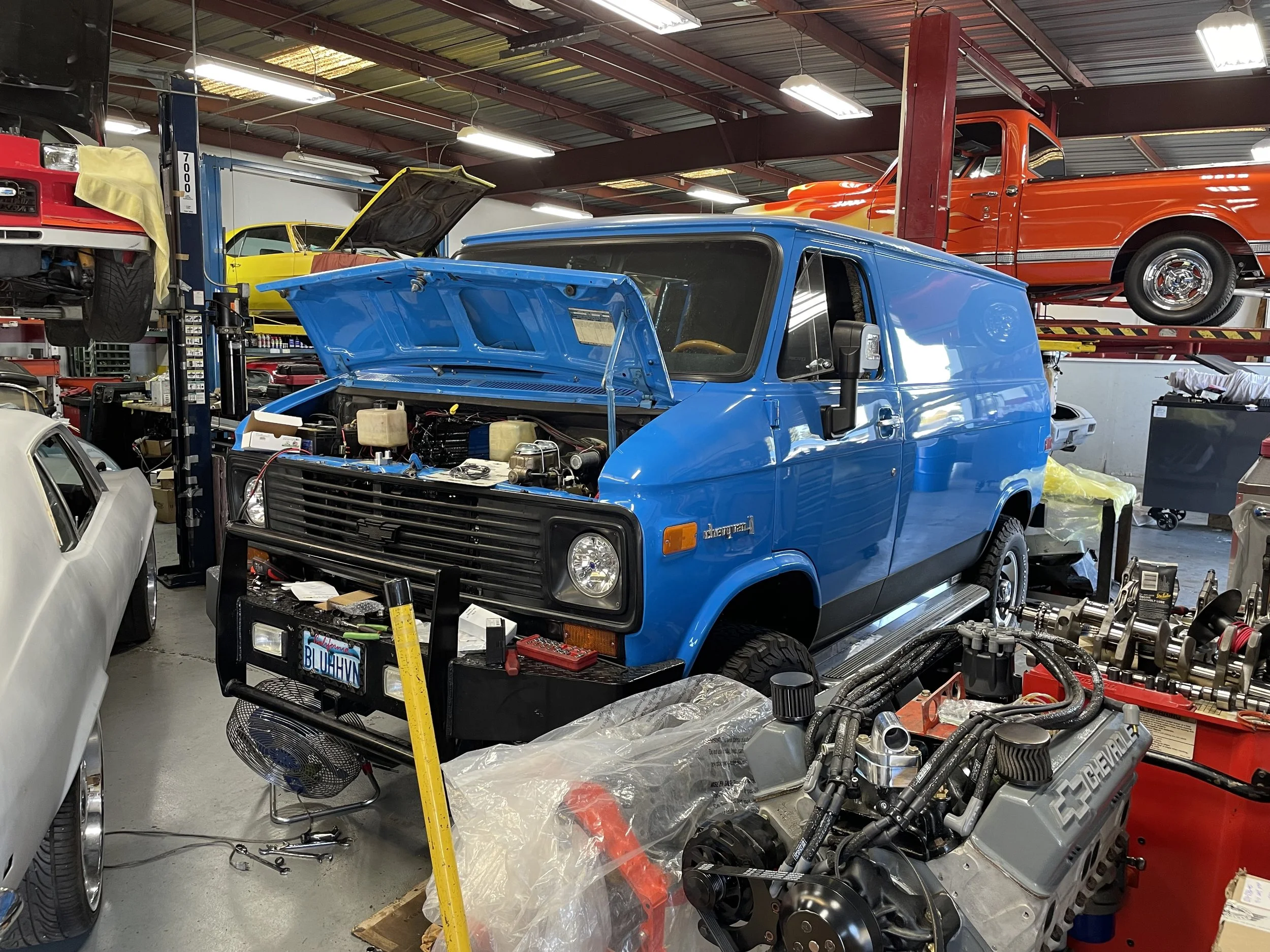 Blue Chevrolet van with open hood in a garage workshop, surrounded by various car parts and tools, with cars on lifts in the background.
