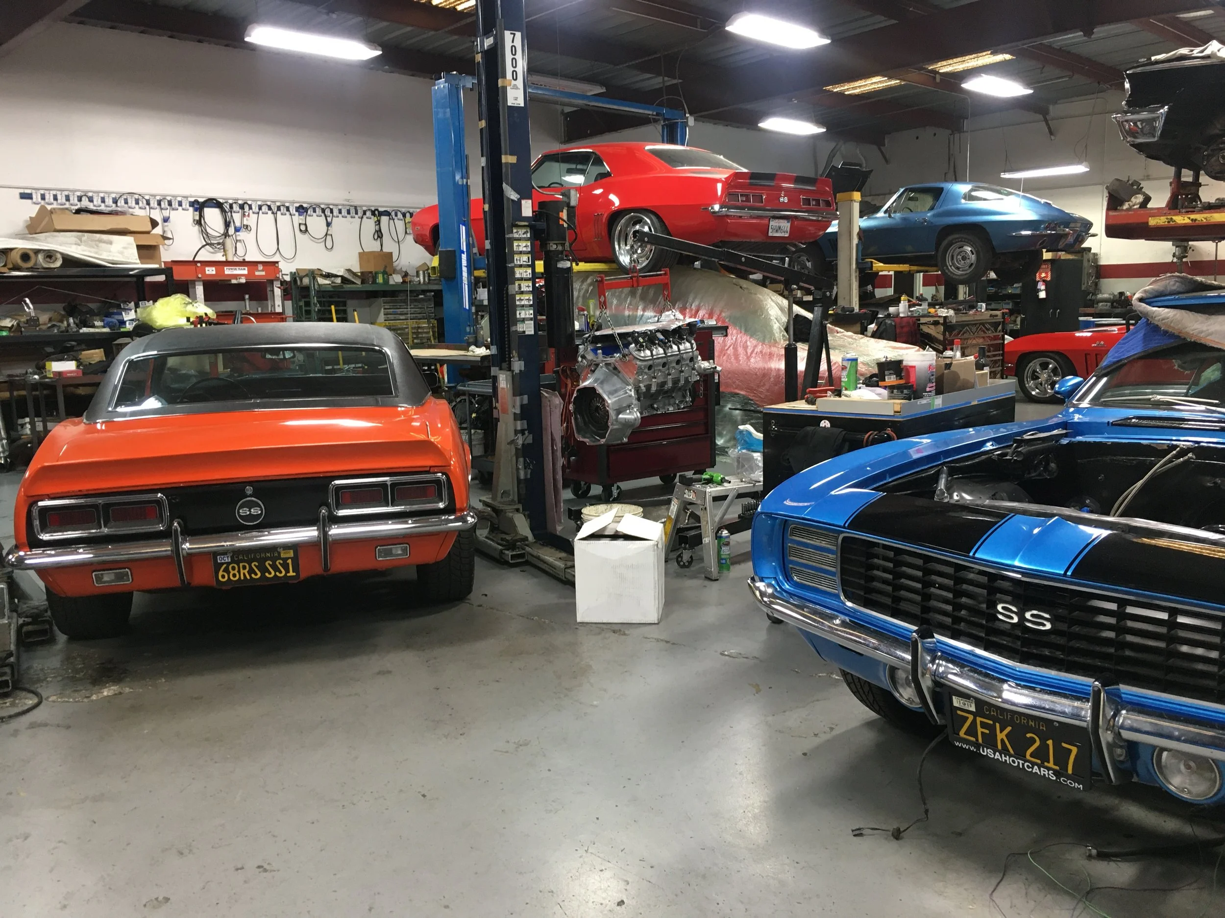 A garage with multiple classic Chevrolet muscle cars in red and blue, some on lifts and others parked on the floor, along with car parts, tools, and equipment scattered around.