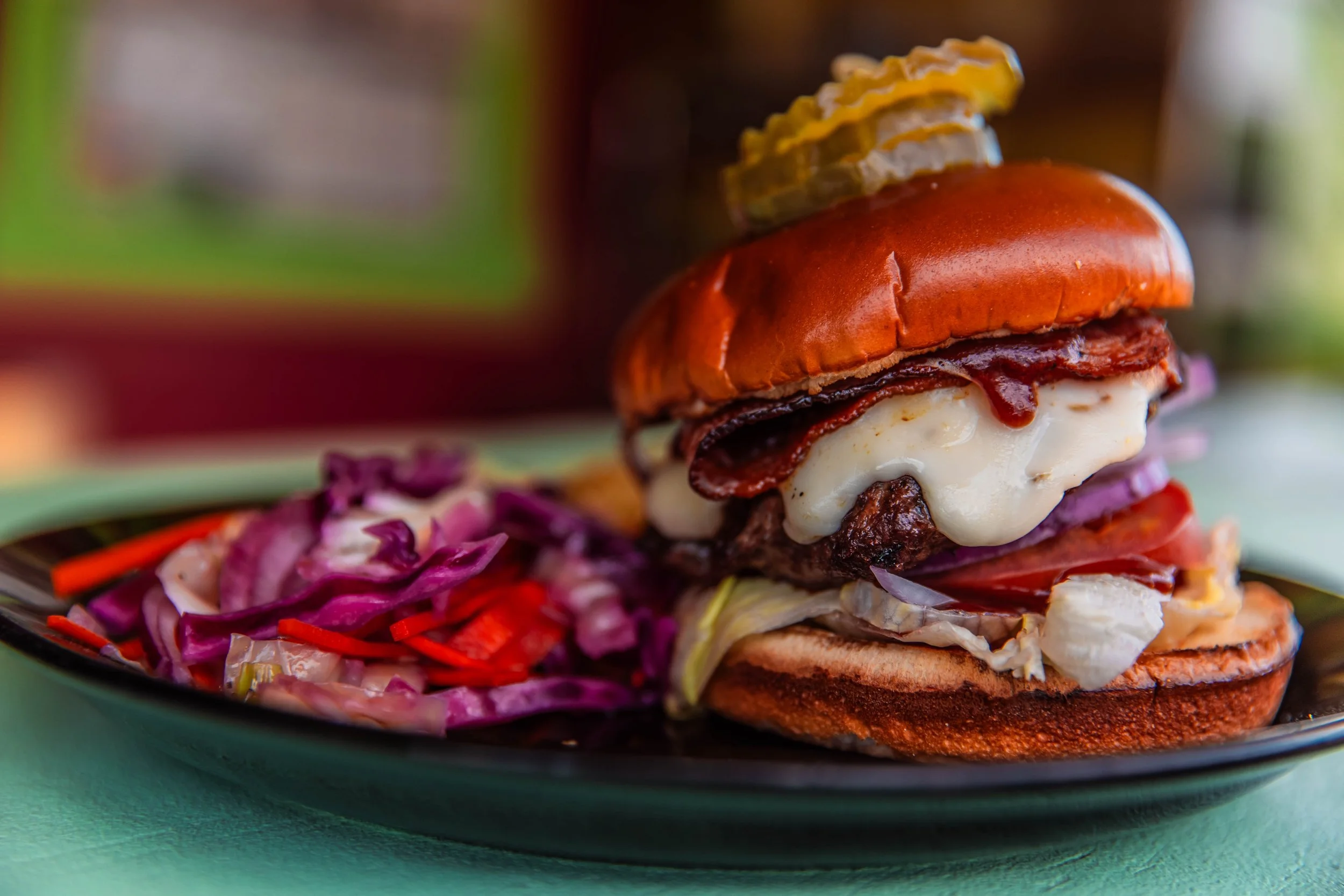 A cheeseburger with bacon, pickles, tomato, lettuce, onions, cheese, and ketchup on a toasted bun, served with coleslaw on a black plate.