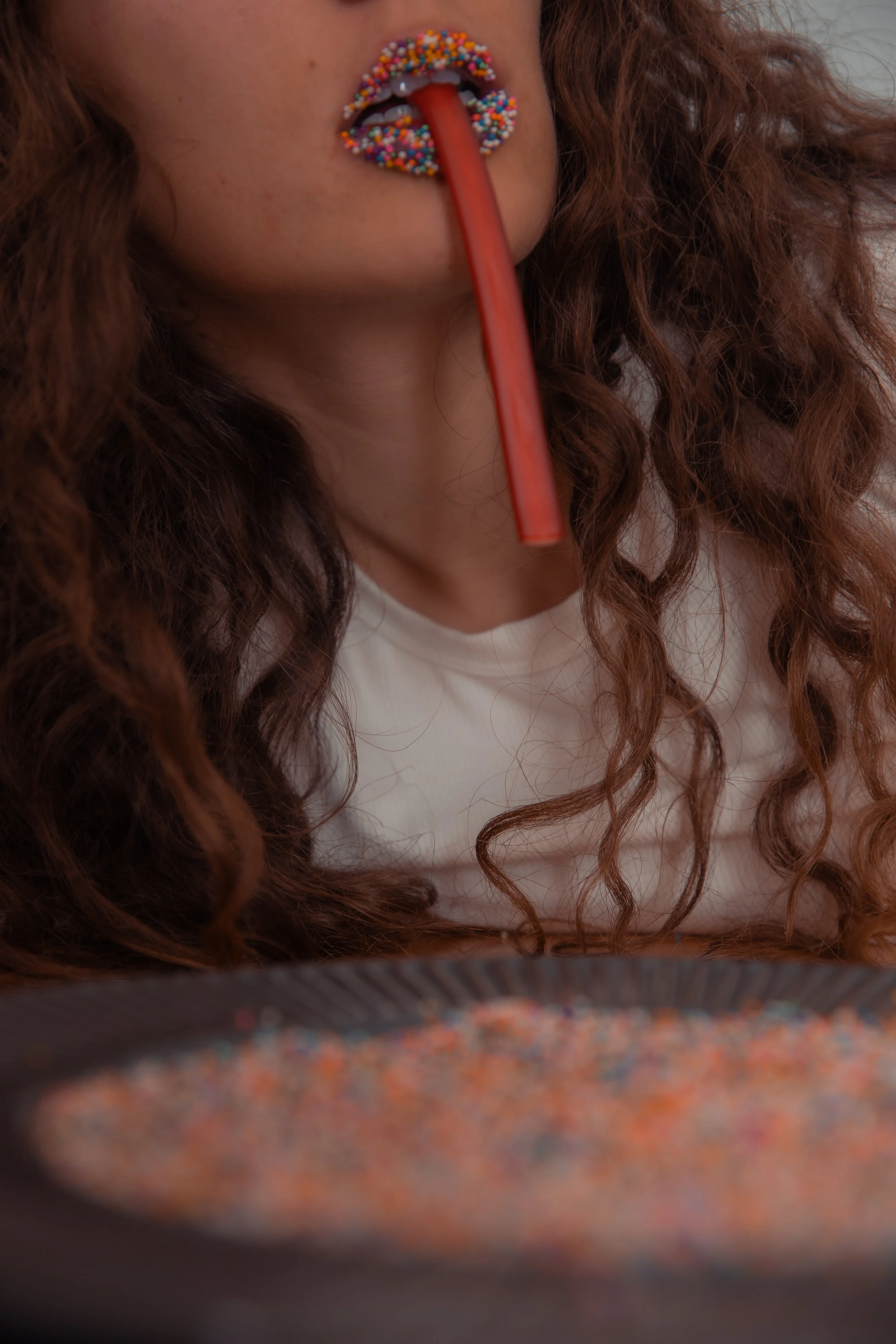 Person with long, curly hair holding a red stick in mouth, lips decorated with colorful sprinkles, and a plate of multicolored sprinkles in front of them.