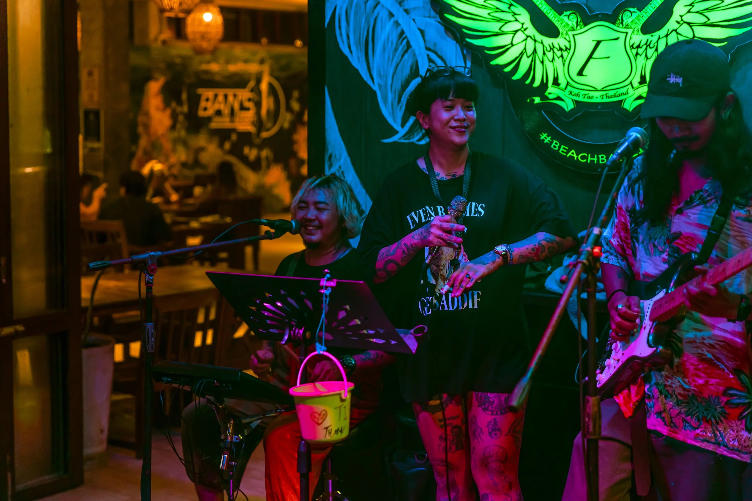 Three musicians performing live; a woman with short blonde hair, a woman with black hair and tattoos holding a microphone, a man wearing a cap playing an electric guitar, in a dimly lit venue with neon signs and a bar in the background.