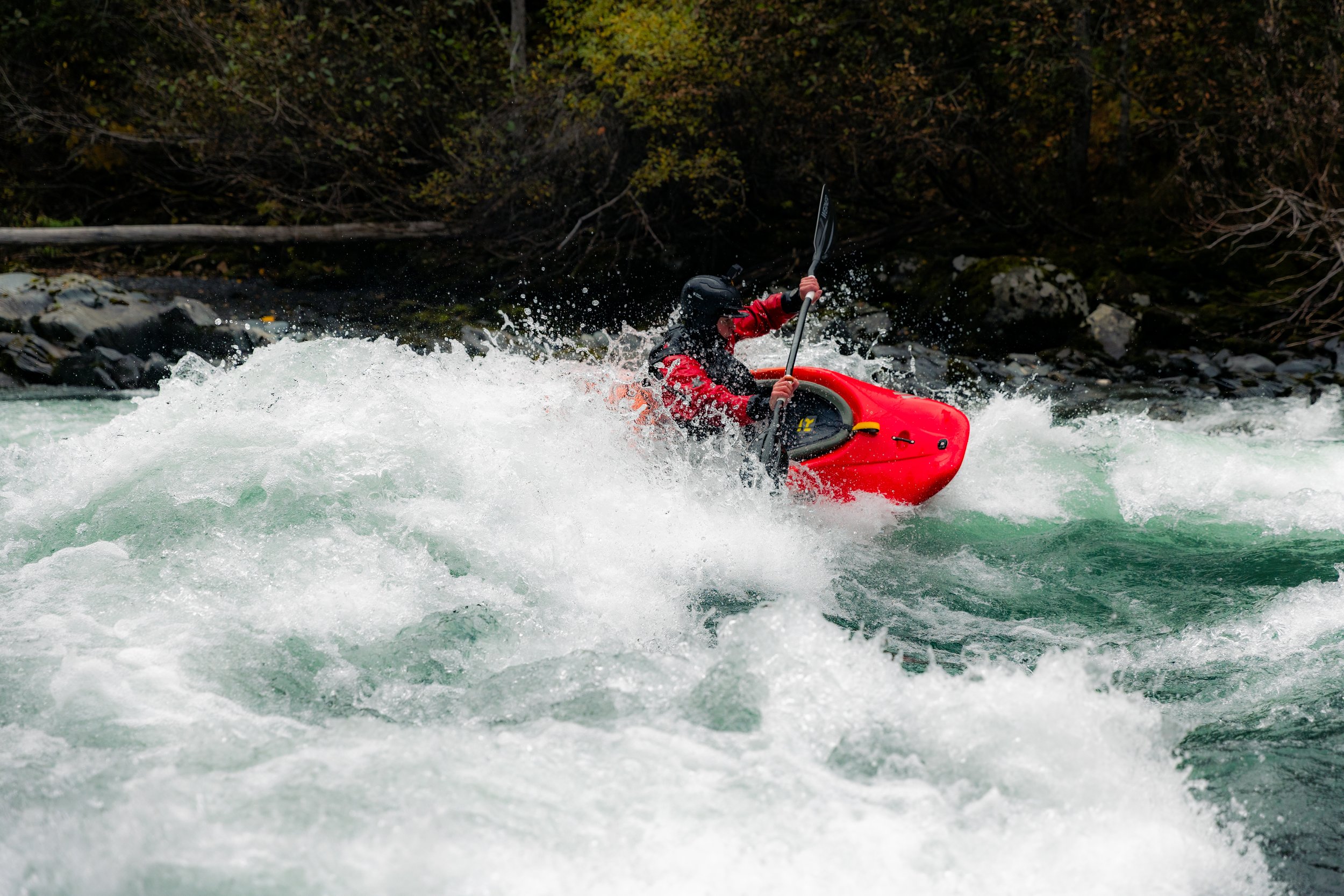Person kayaking in rushing white water river with splash and waves, wearing helmet and red waterproof gear.