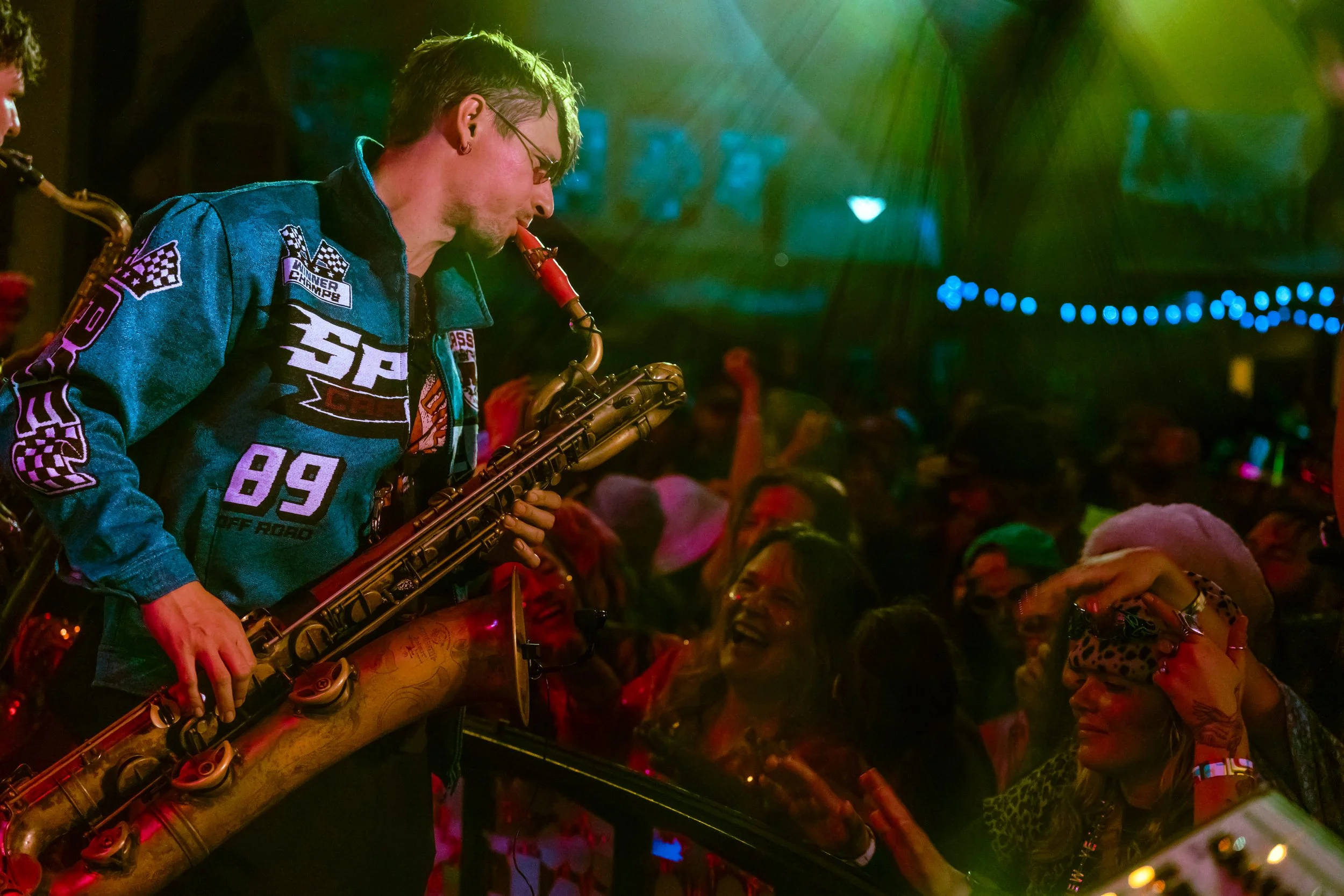 A musician playing a saxophone at a lively, colorful party. The crowd is dancing and smiling, with vibrant lights and decorations in the background.