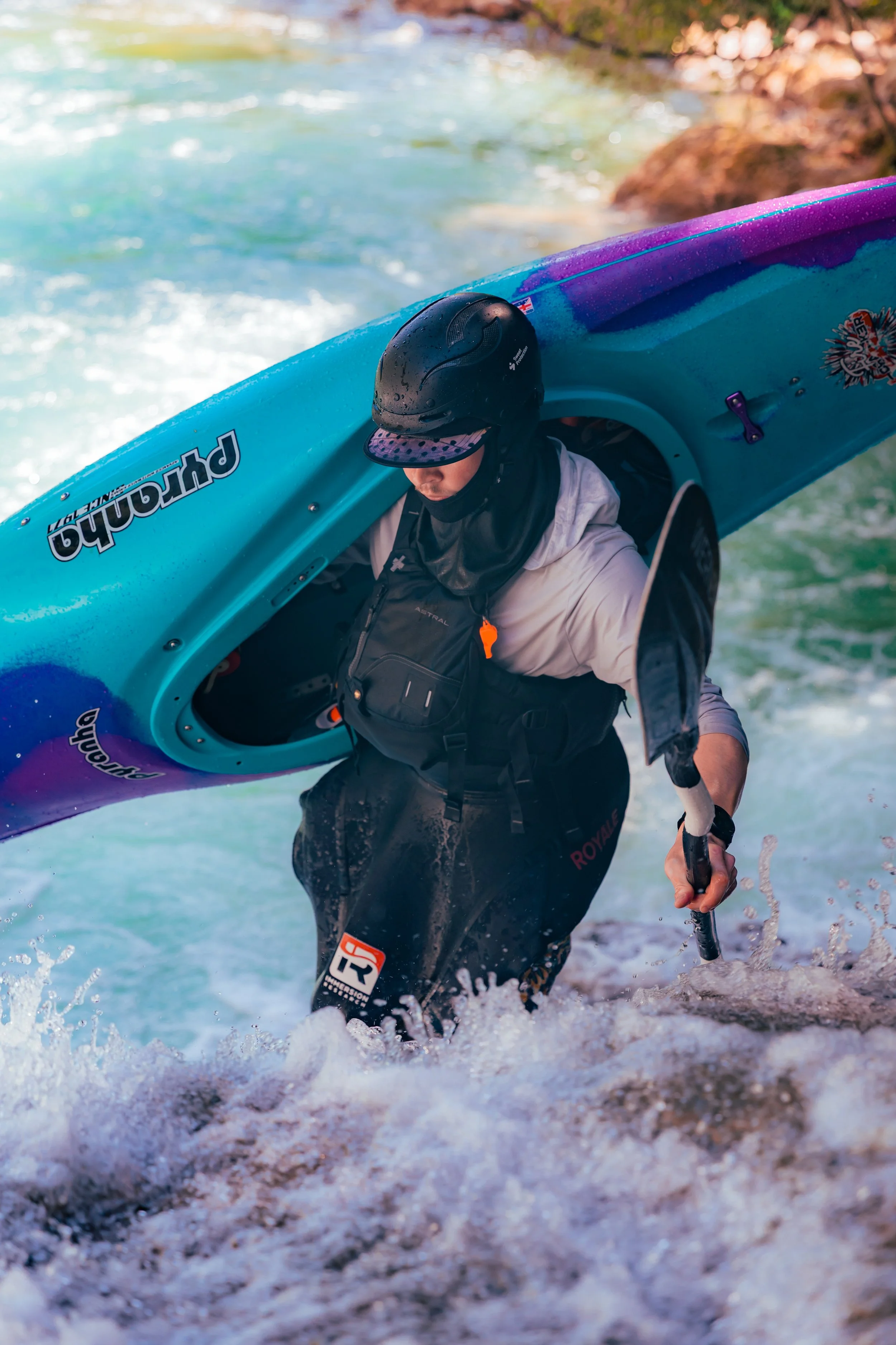 Person in a black helmet and life vest emerging from turbulent river with a colorful kayak.