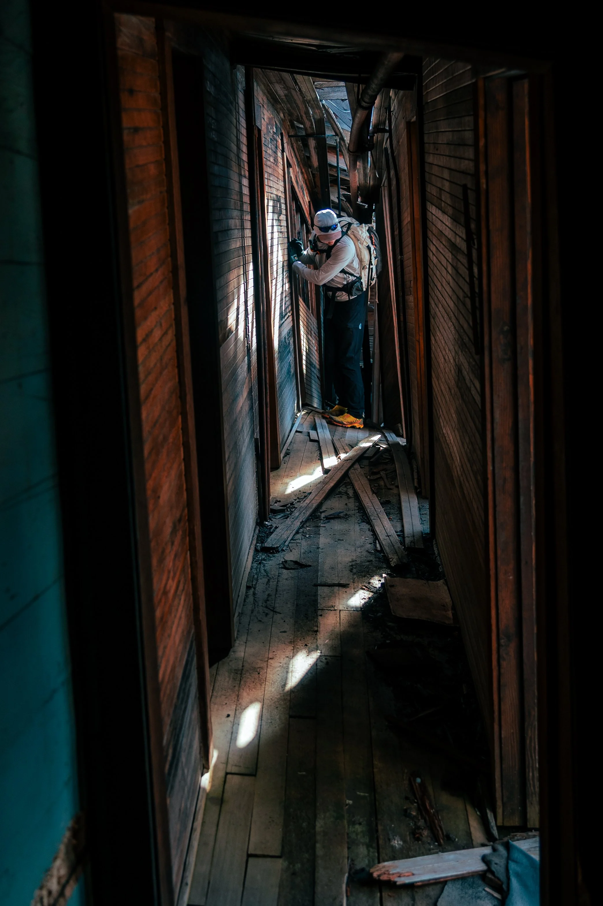 A person wearing a white helmet and outdoor gear inspecting a damaged and charred wooden interior hallway with damaged walls and scattered debris.