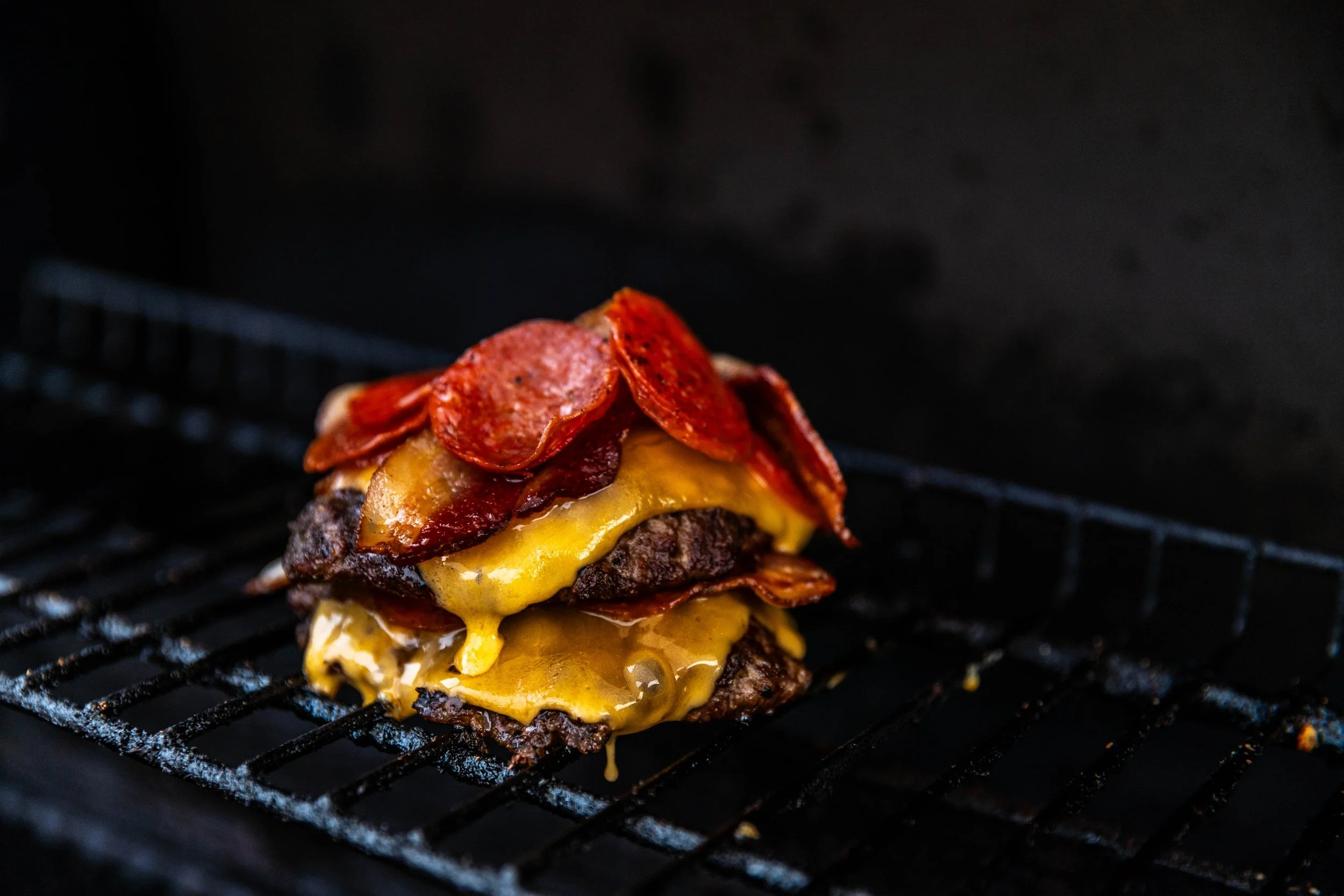 Close-up of a cheeseburger with bacon and pepperoni slices on a grill rack in a black background.