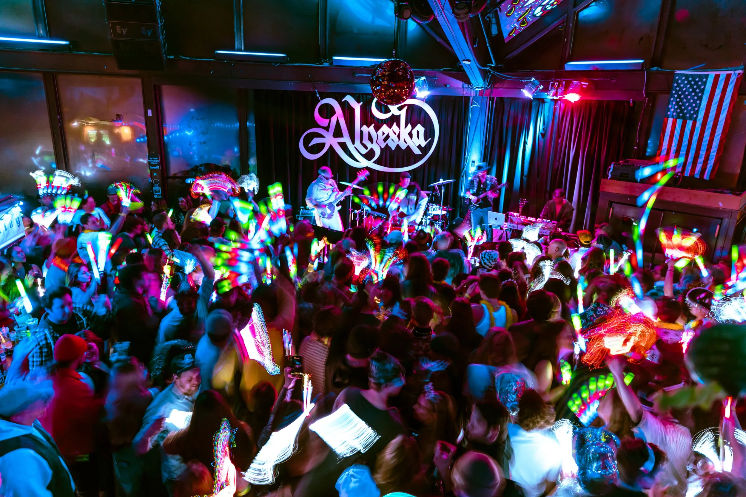 A lively concert at Alyeska with a band playing on stage and a crowd dancing with colorful glow sticks in a night club setting.