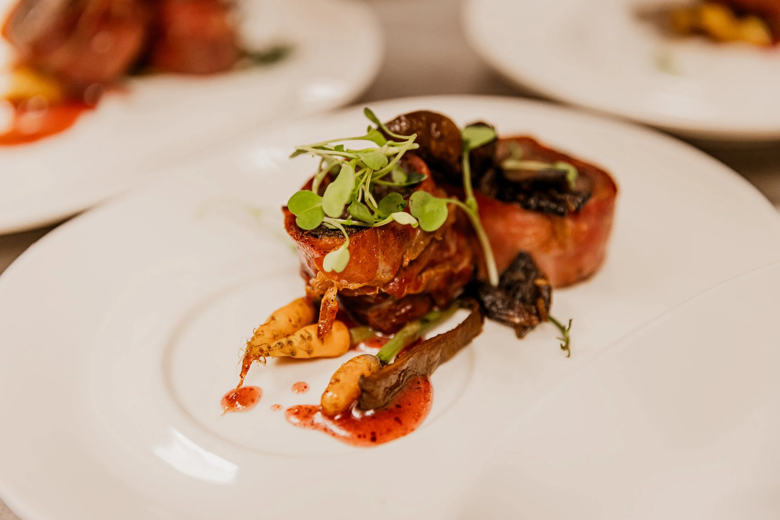 A plated dish featuring a piece of steak topped with microgreens, accompanied by roasted vegetables and a red sauce.