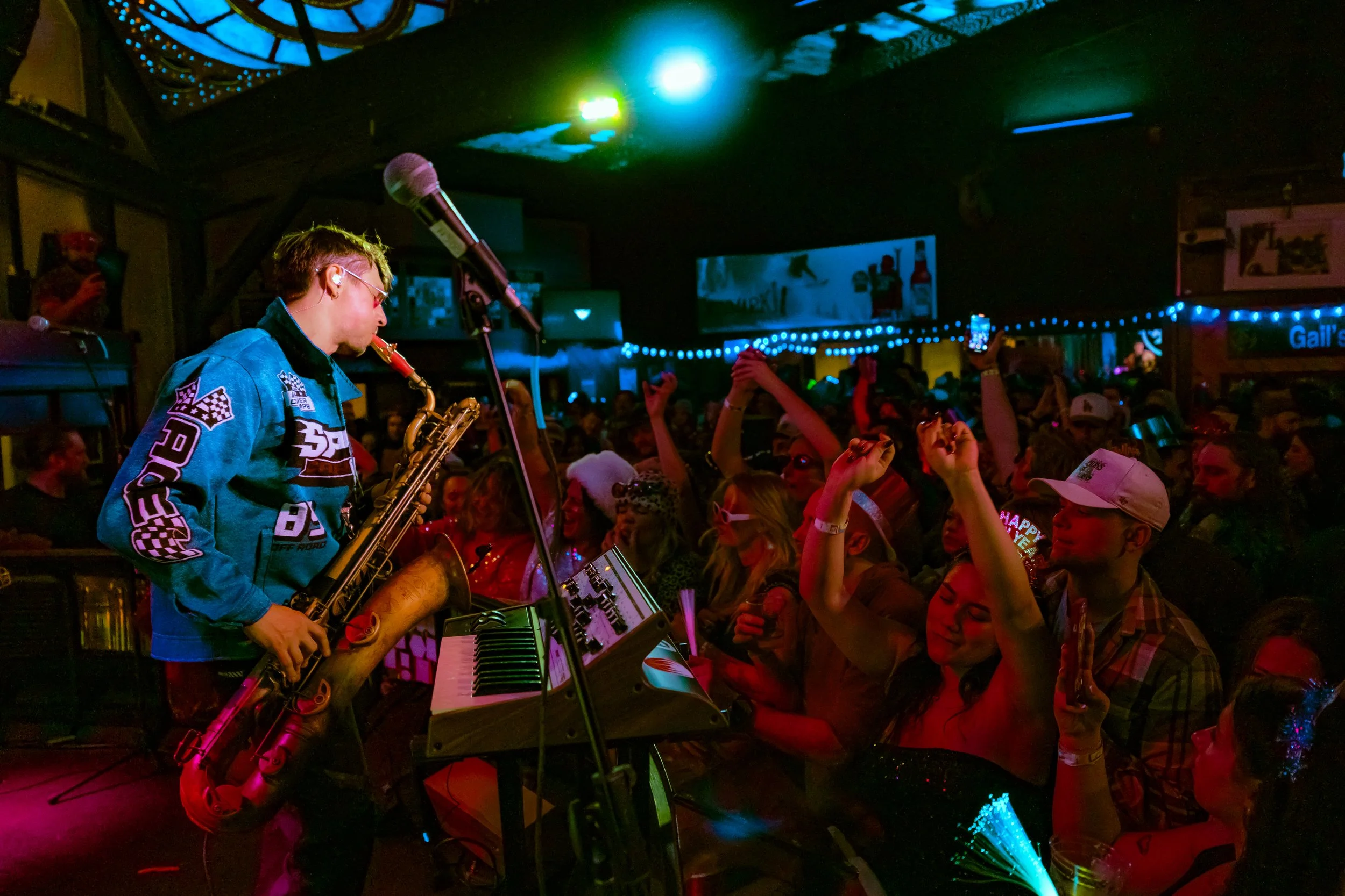 A musician playing the saxophone on stage in a lively indoor concert with an excited crowd dancing and raising their hands.