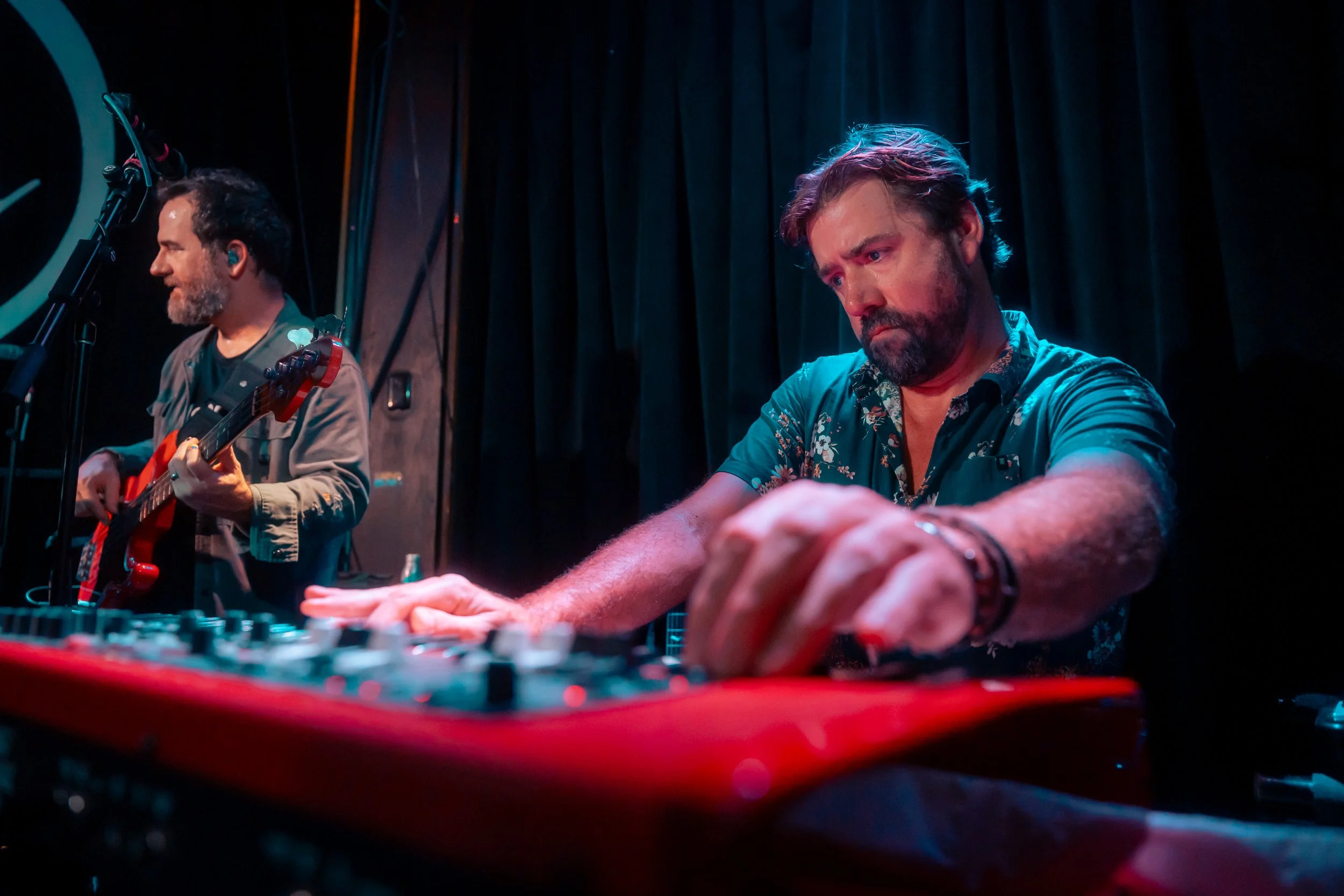 Two male musicians perform on stage; one plays a guitar, and the other adjusts sound equipment, with black curtains in the background.