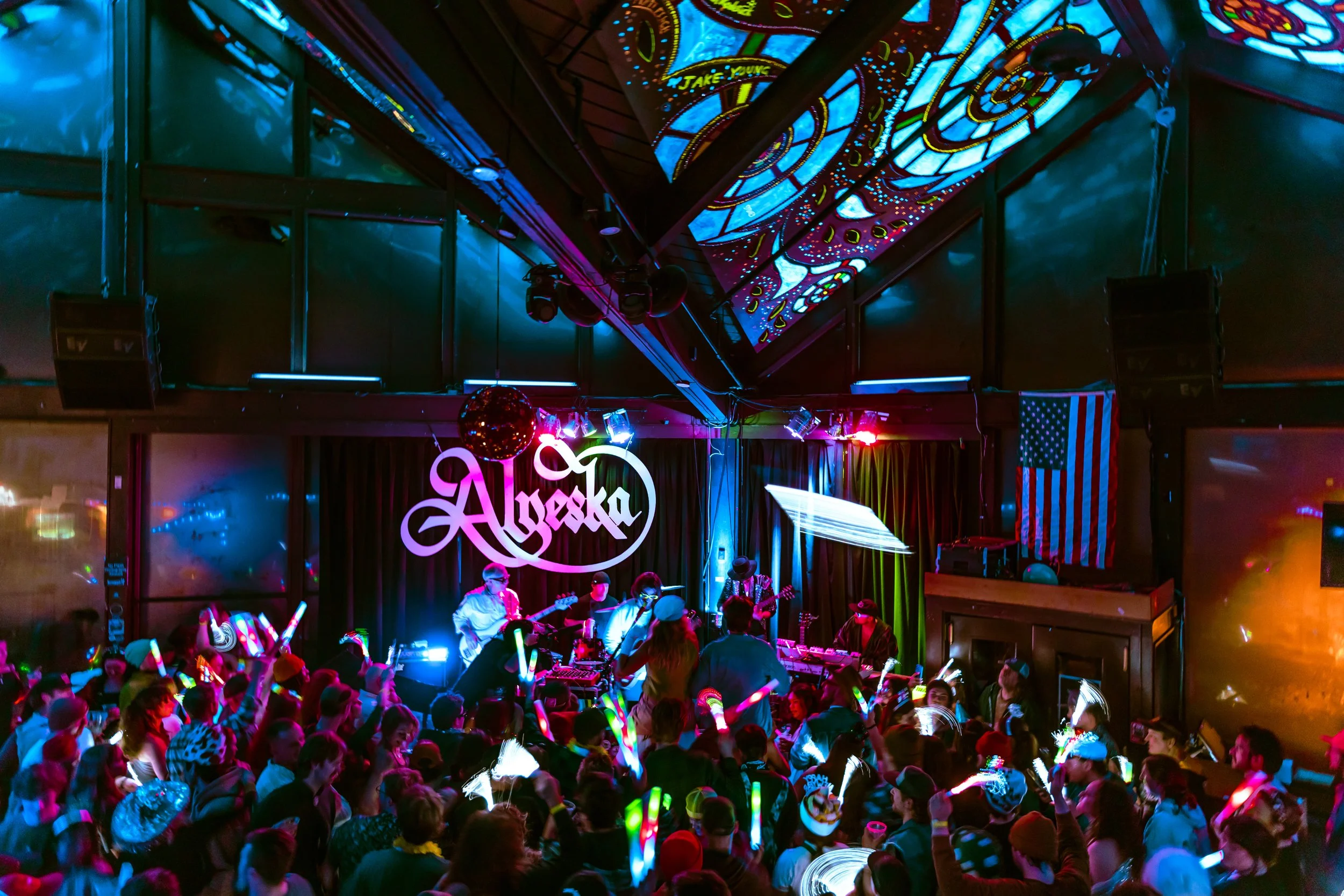 A lively indoor concert with a band playing on stage for a crowd of people holding glow sticks. The stage has a large neon sign reading 'Alaska', colorful lights, and an American flag in the background. The ceiling is decorated with vibrant, patterne