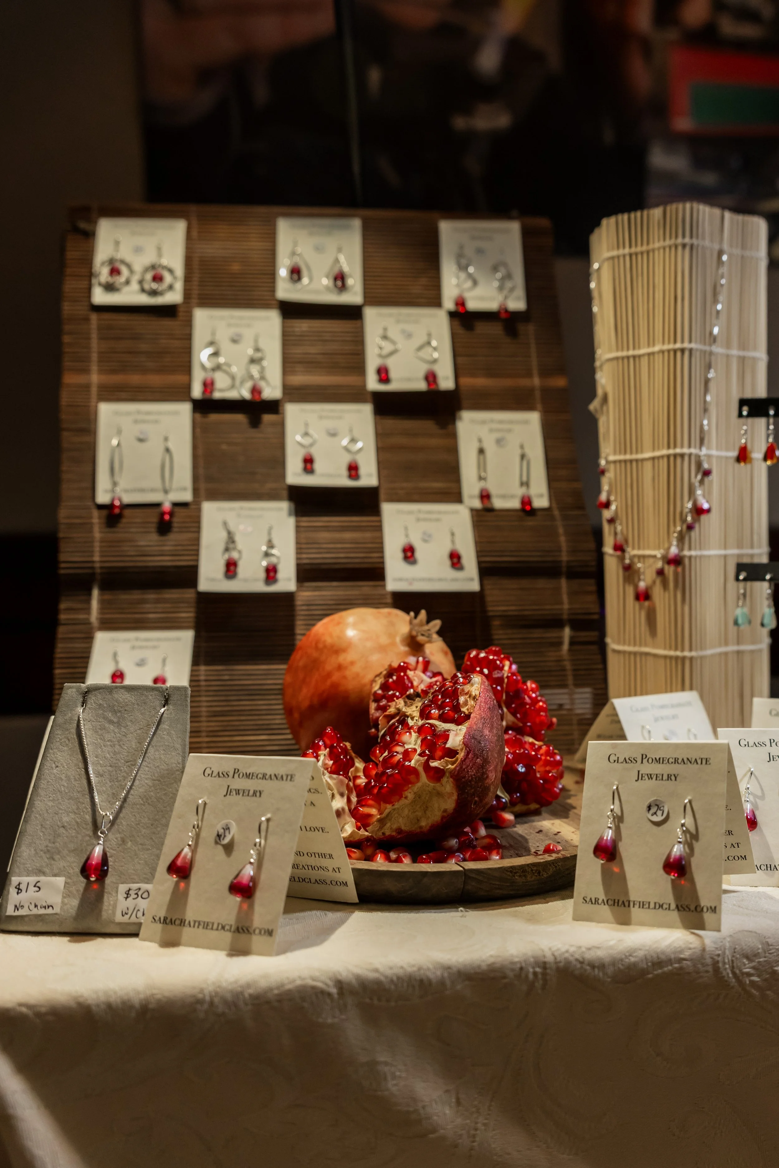 Display of jewelry with pomegranate theme, including necklaces and earrings, arranged beside a pomegranate. Jewelry tags show prices and website information. The background features a wooden stand with more jewelry and seedless pomegranate beads hang