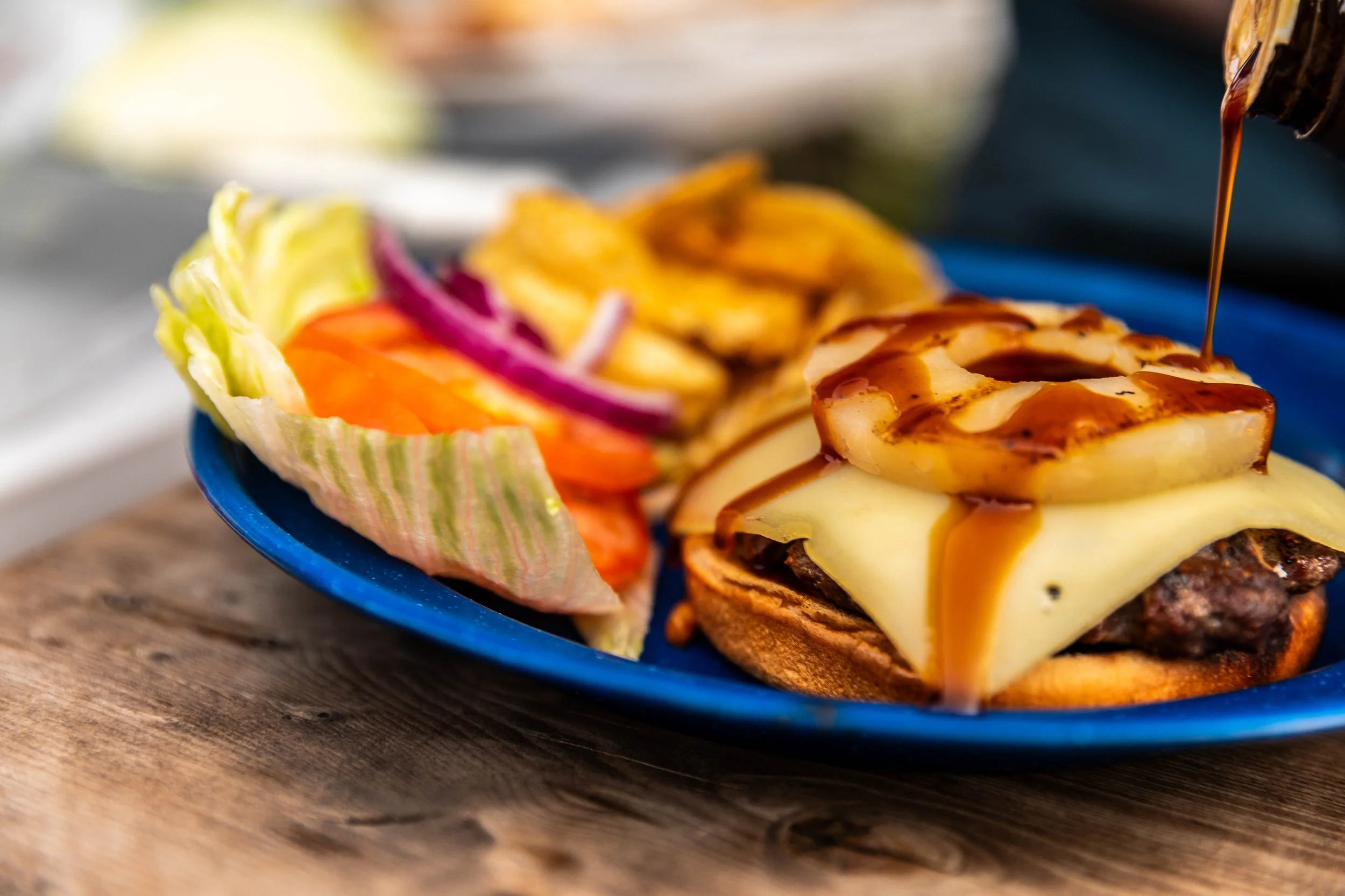 Cheeseburger with grilled onions and barbecue sauce, served with fresh vegetables and potato chips on a blue plate.