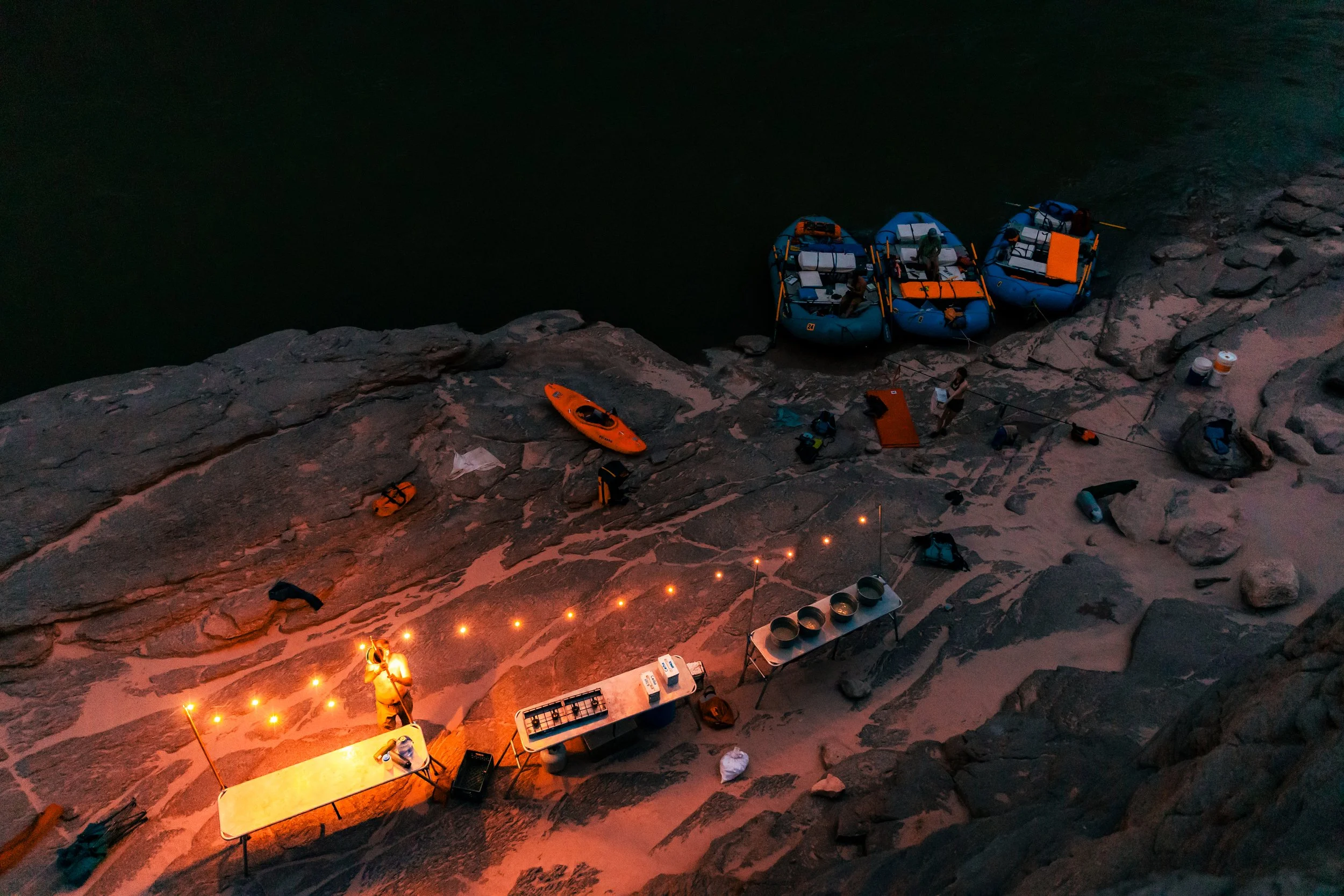 Aerial view of a daytime outdoor rafting campsite on rocky terrain beside a river, with three blue inflatable rafts, orange kayaks, camping gear, and a makeshift table with supplies.