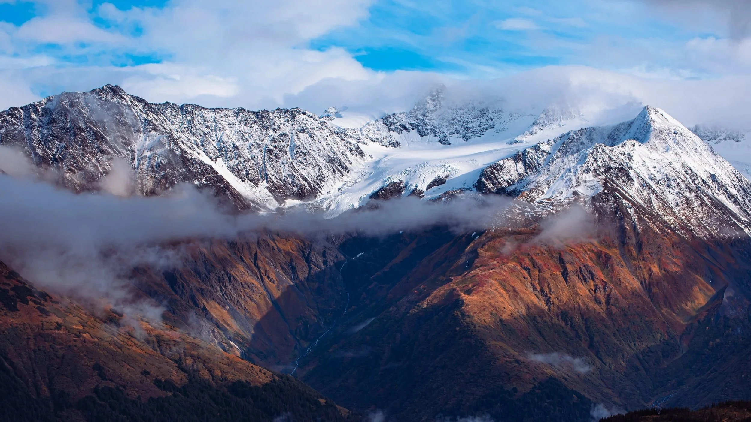 Snow-capped mountains with clouds and blue sky, steep rugged slopes with autumn-colored vegetation.