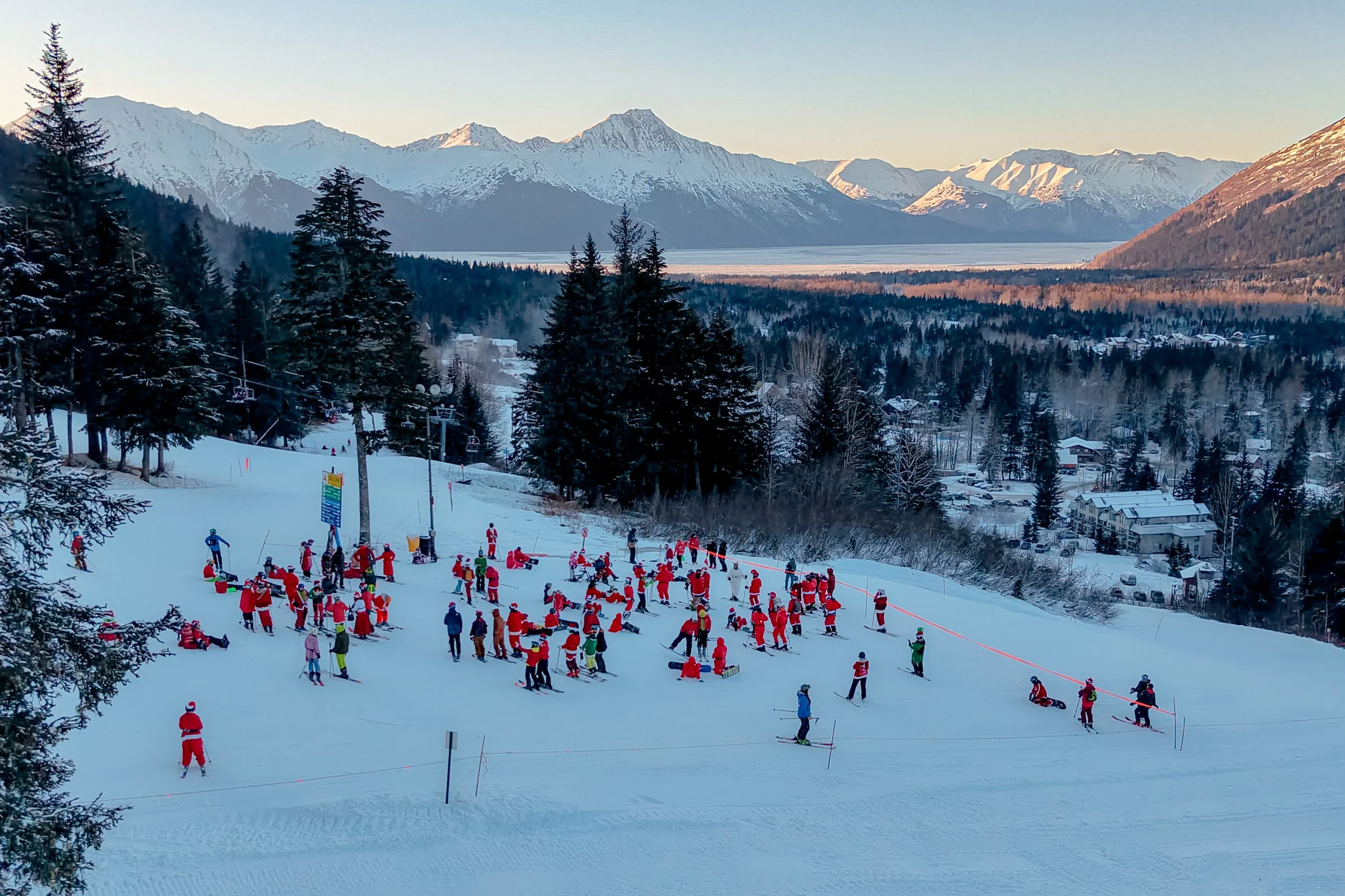 A snowy ski resort with many people dressed in Santa costumes, skiing and snowboarding, set against a backdrop of snowy mountains and a lake.