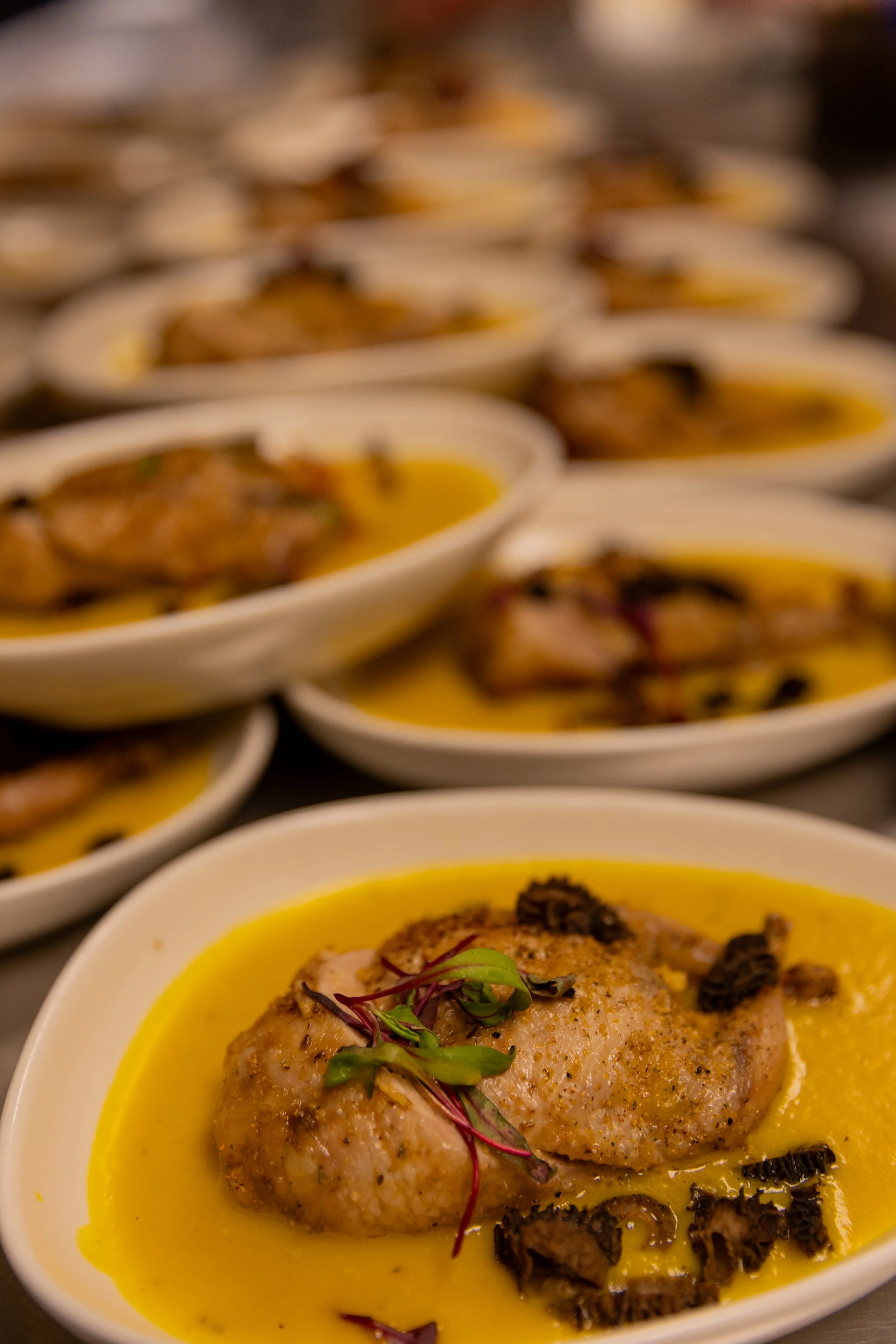 Close-up of a dish with a piece of cooked chicken on yellow sauce, garnished with herbs and black truffle pieces, arranged in bowls on a table.