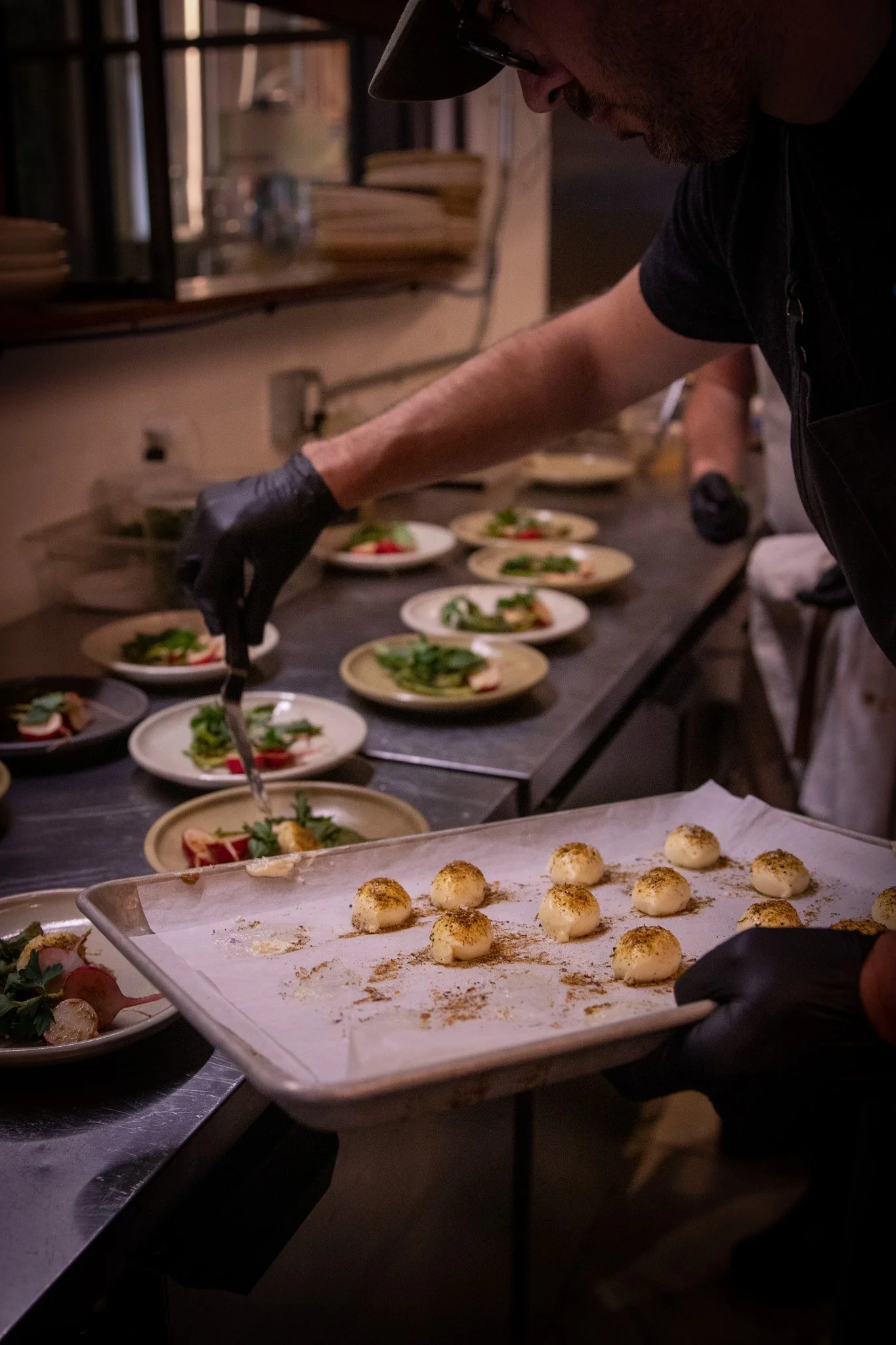 Chef preparing multiple plates of food, including small round appetizers on a tray, in a professional kitchen.