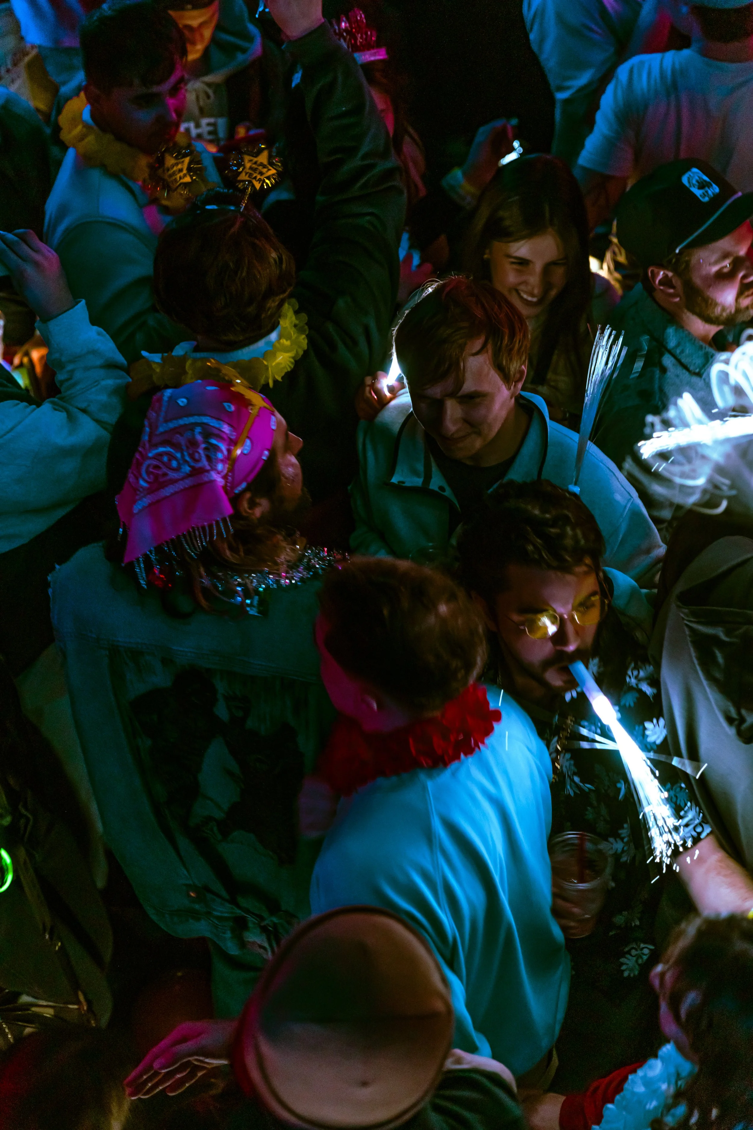 A crowded scene with people celebrating a party or event, some wearing festive accessories like leis, hats, and headbands, with colorful lighting and sparklers.