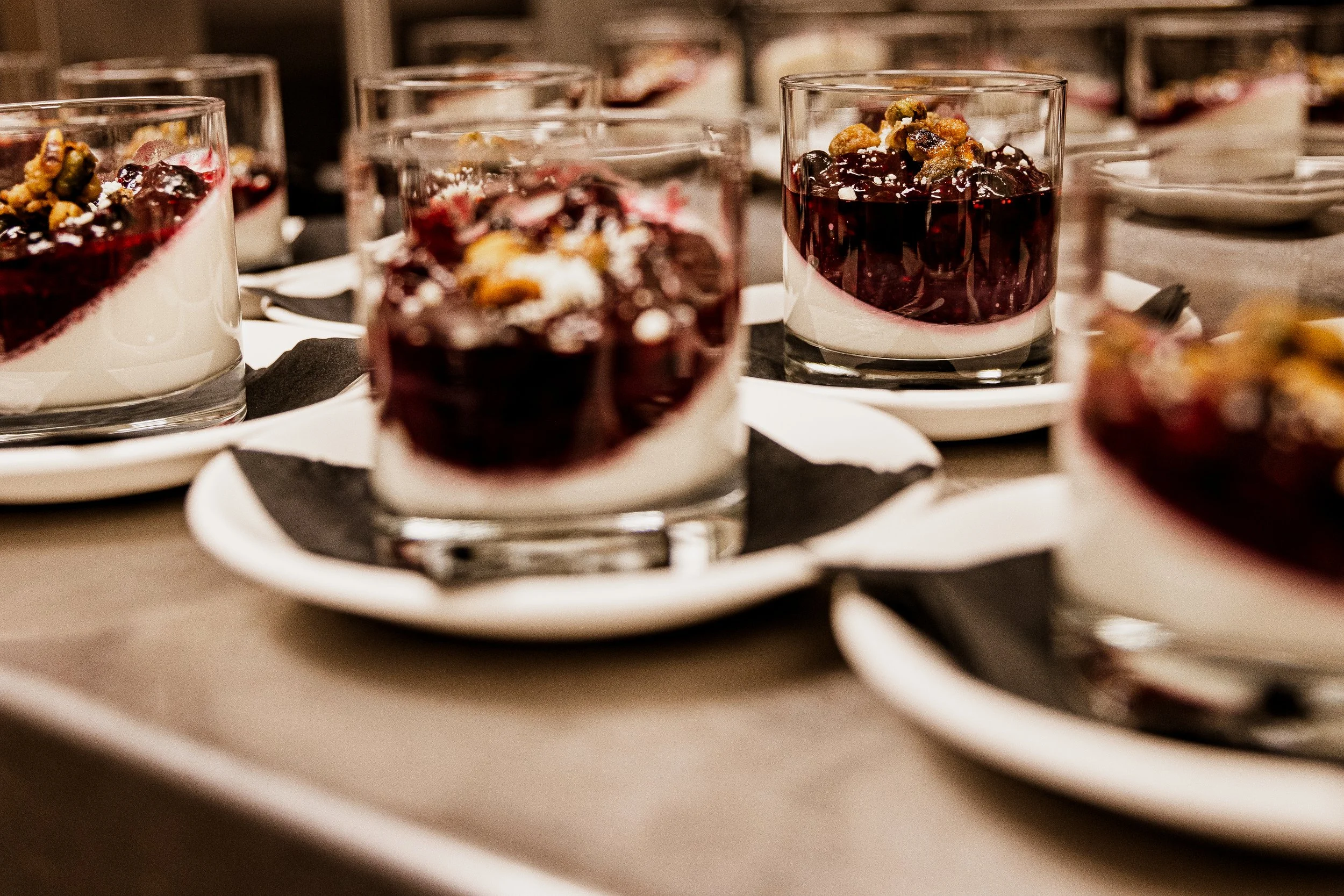 Close-up of layered dessert cups with dark berry topping, white cream layer, served in clear glasses on white plates with black napkins, set on a buffet table.