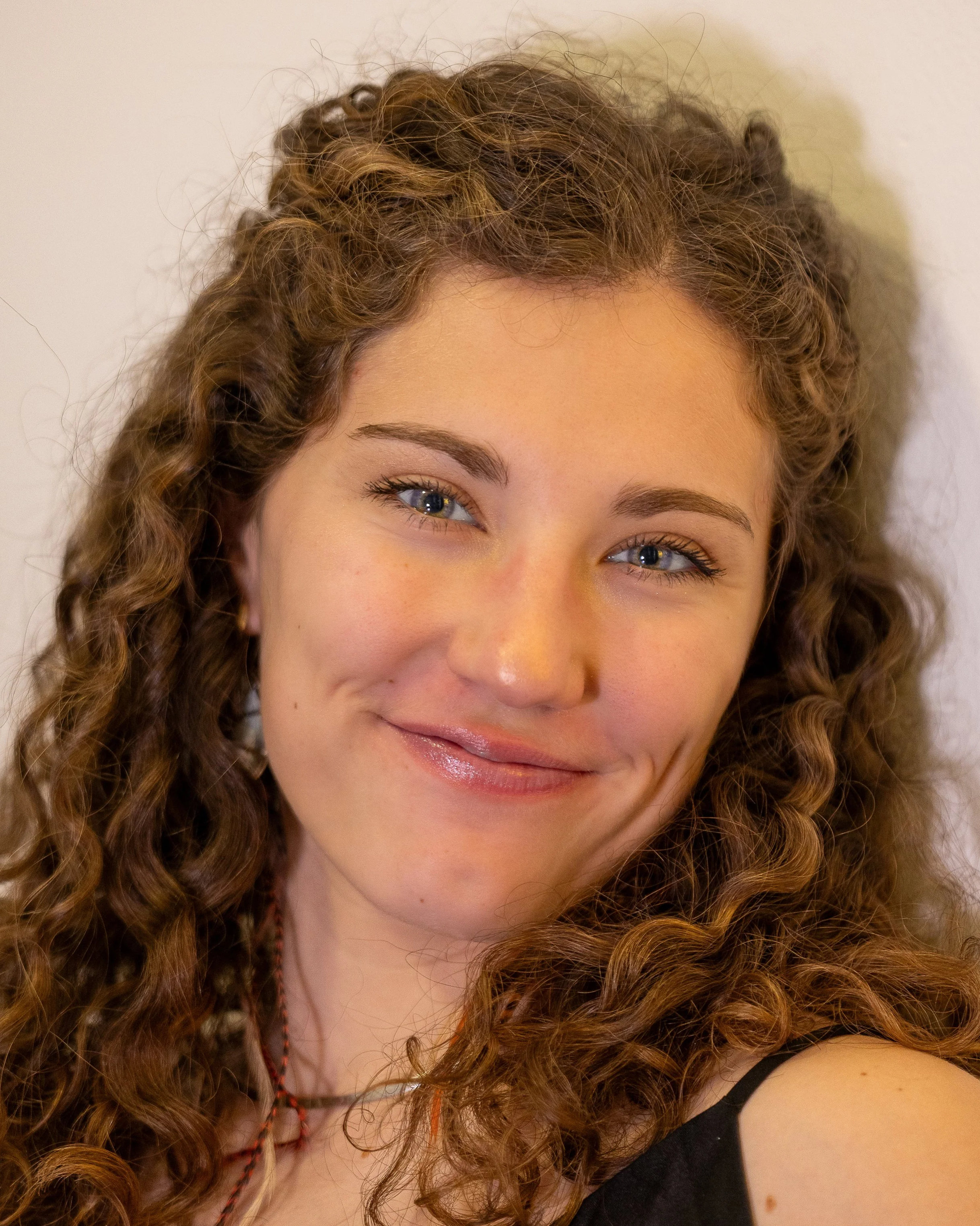 Close-up of a smiling woman with curly hair, blue eyes, and wearing a black top and colored beaded necklace.