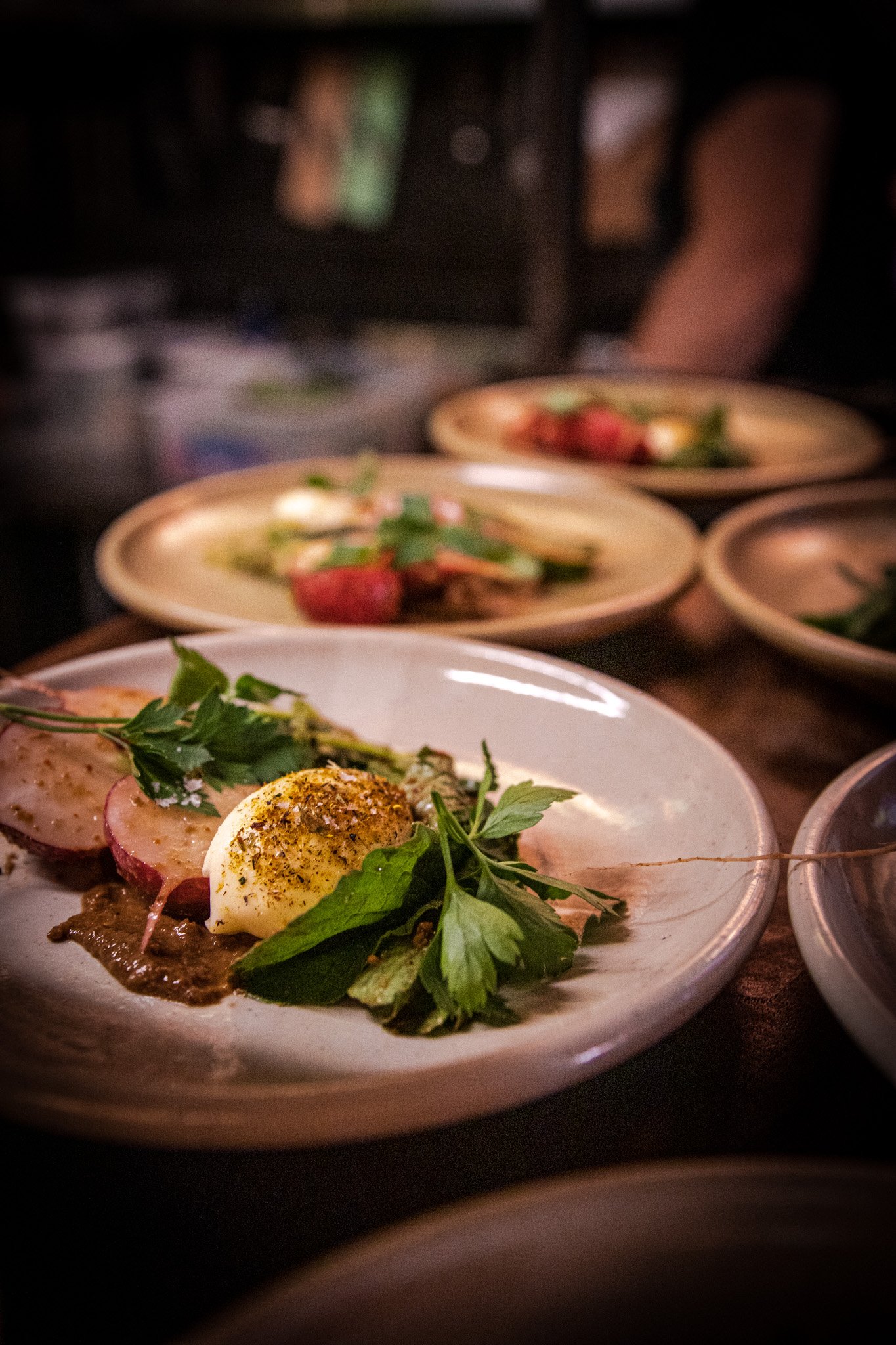 Close-up of a plate with a soft-boiled egg topped with spices, parsley, and slices of radish, with other plates of food blurred in the background.