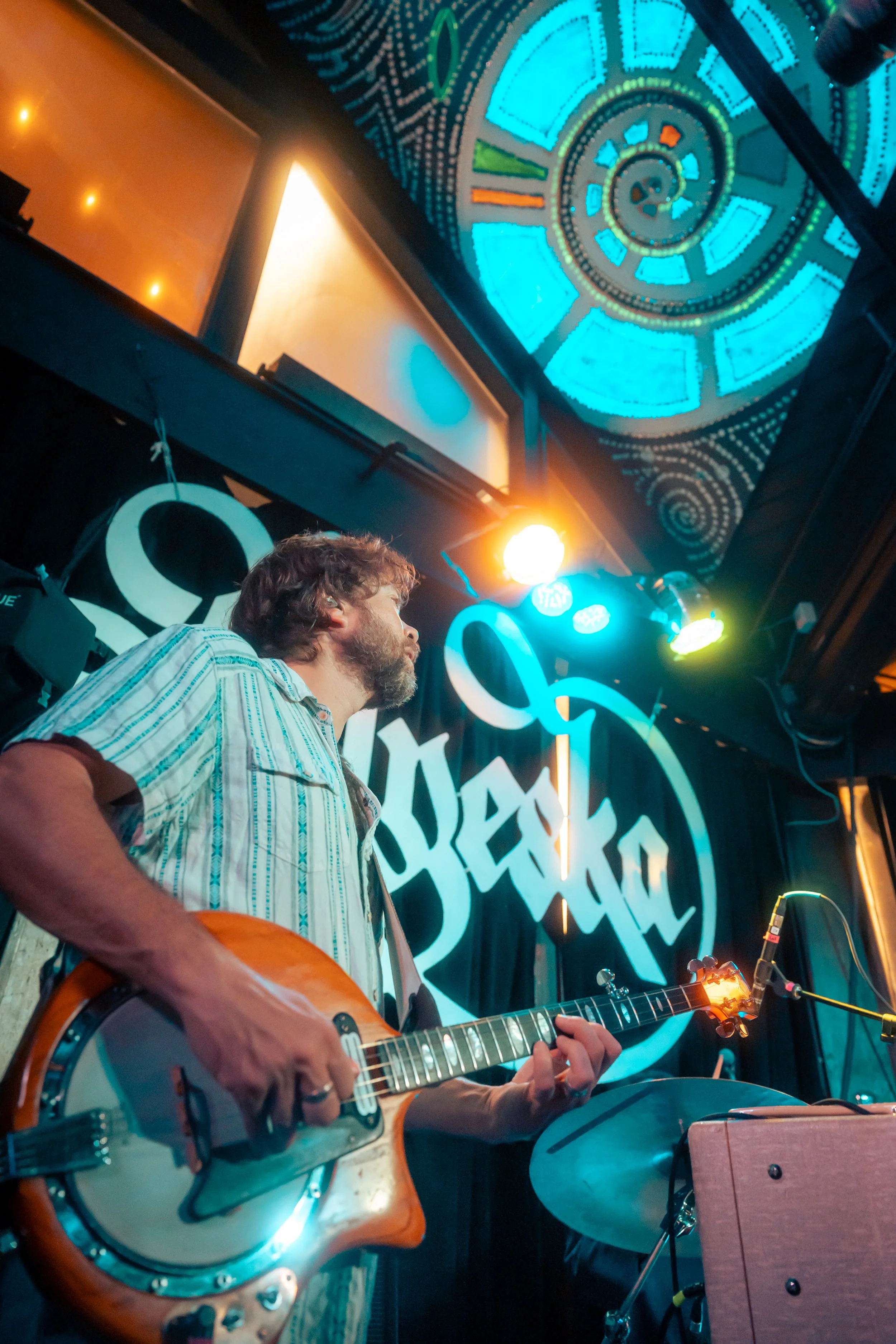 A male musician playing a guitar on stage at a venue named 'Sexto.' The background features a black curtain with the logo and neon lights, as well as a decorative ceiling with colorful geometric patterns.
