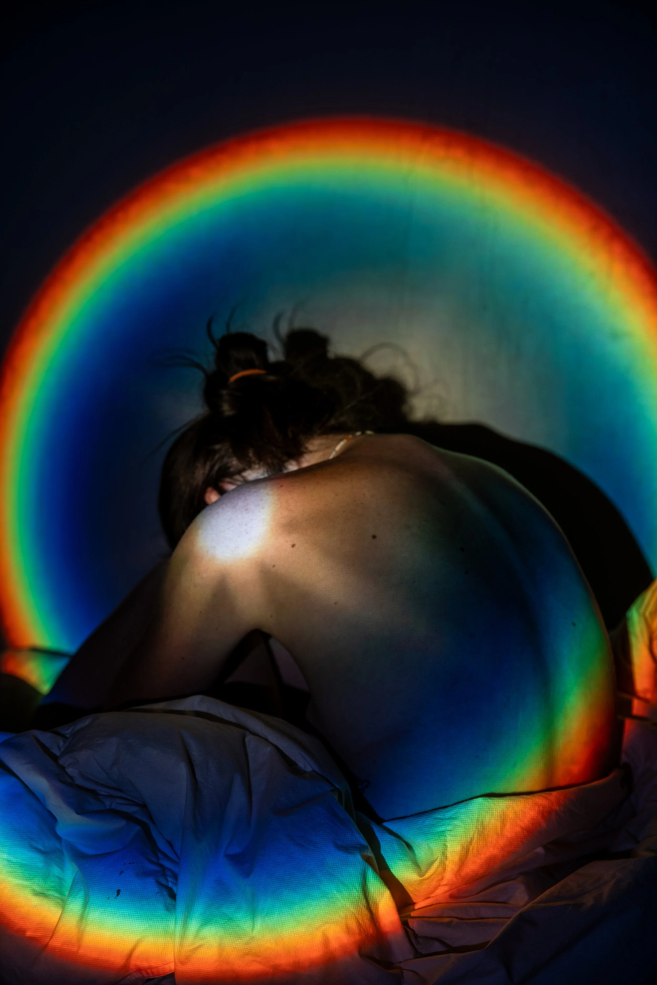 Person sitting with their back to the camera, surrounded by colorful rainbow light projections on a dark background.