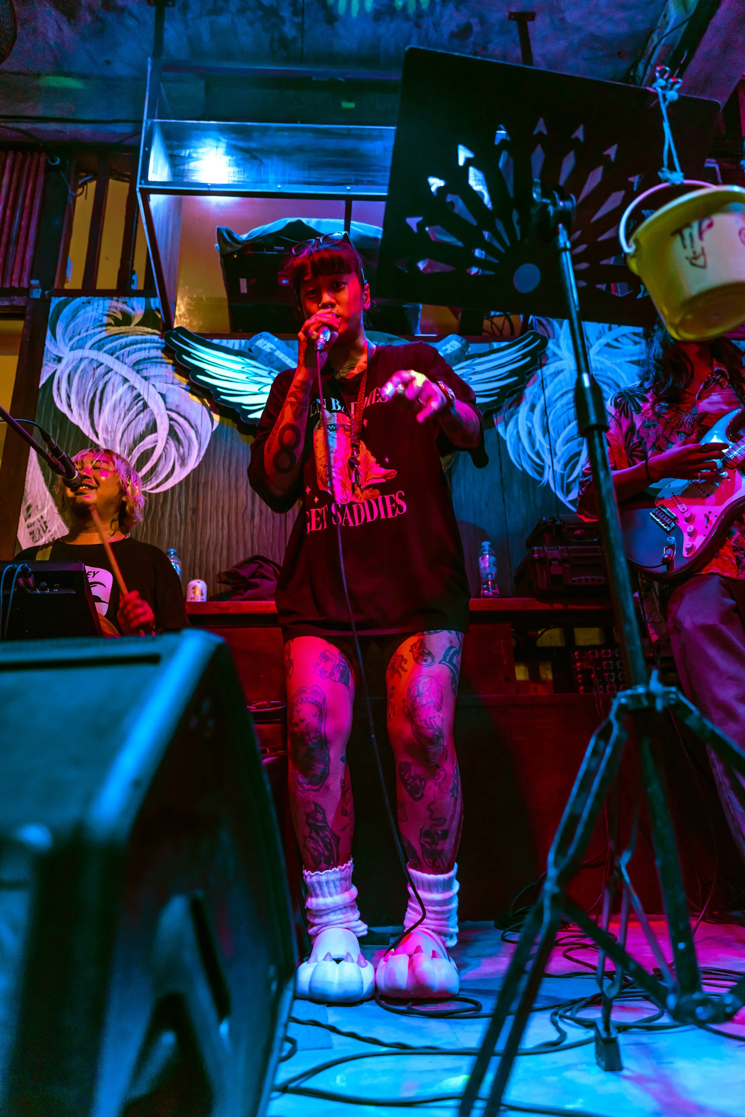 A female performer with tattoos on her legs singing into a microphone on a colorful stage, with a male musician playing drums on her left and a guitarist on her right, under vibrant stage lighting.
