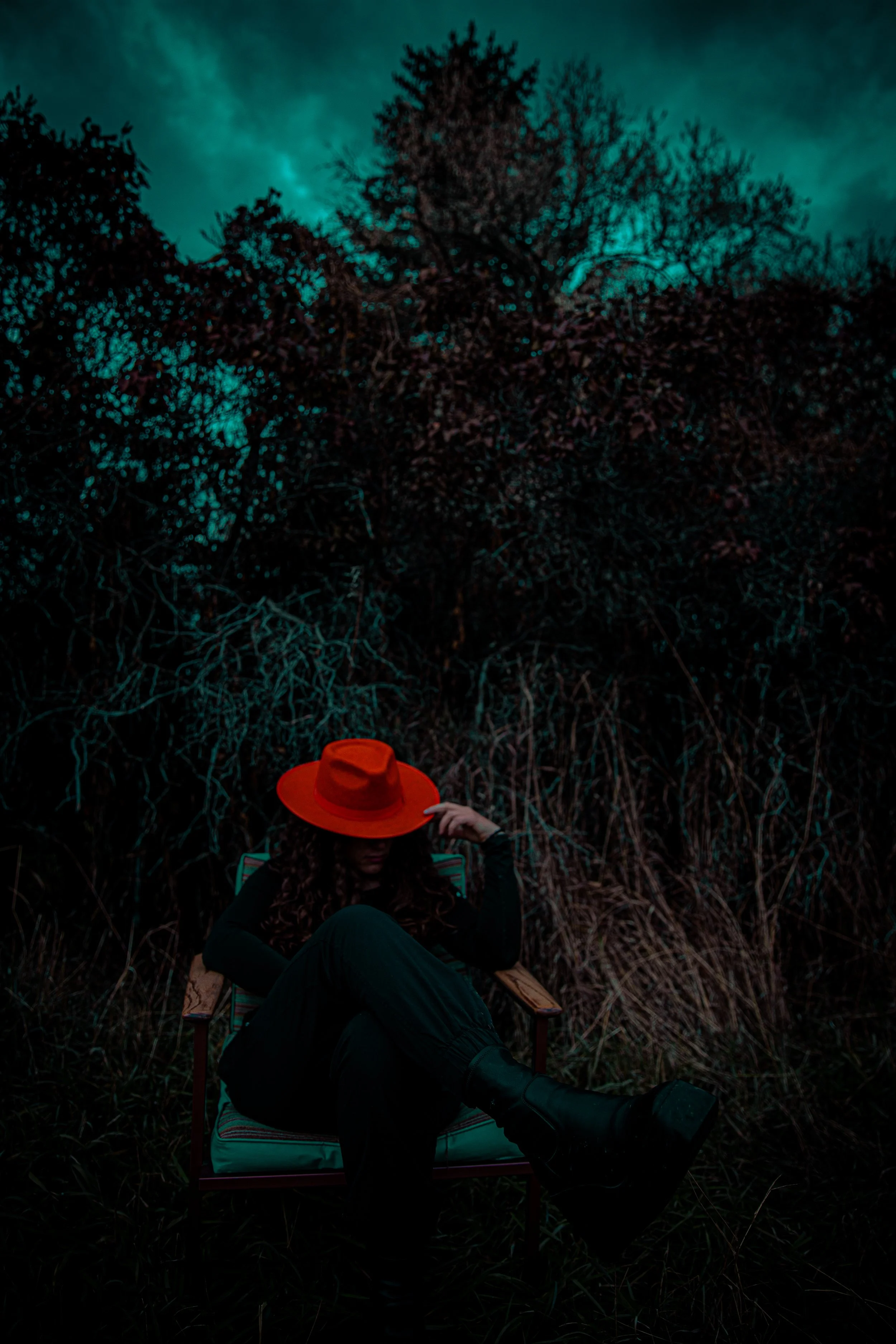 A woman sitting on a chair outdoors at dusk, wearing a large red hat, black outfit, and black boots, with tall grass and trees in the background and a dark, cloudy sky.