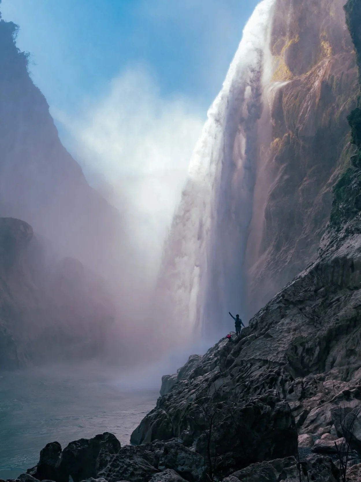 A person standing on rocks near a waterfall, raising one arm, with mist and cliffs surrounding the waterfall.