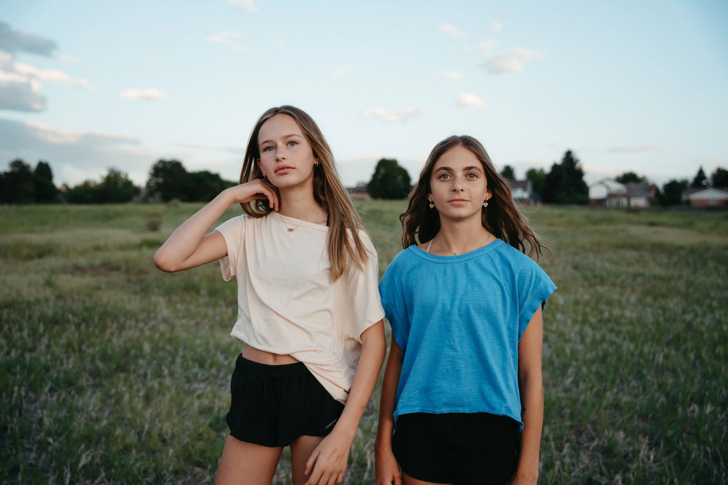 Two young women standing outdoors in a grassy field with trees and houses in the background, one wearing a beige t-shirt and black shorts, the other wearing a blue t-shirt, both looking at the camera.