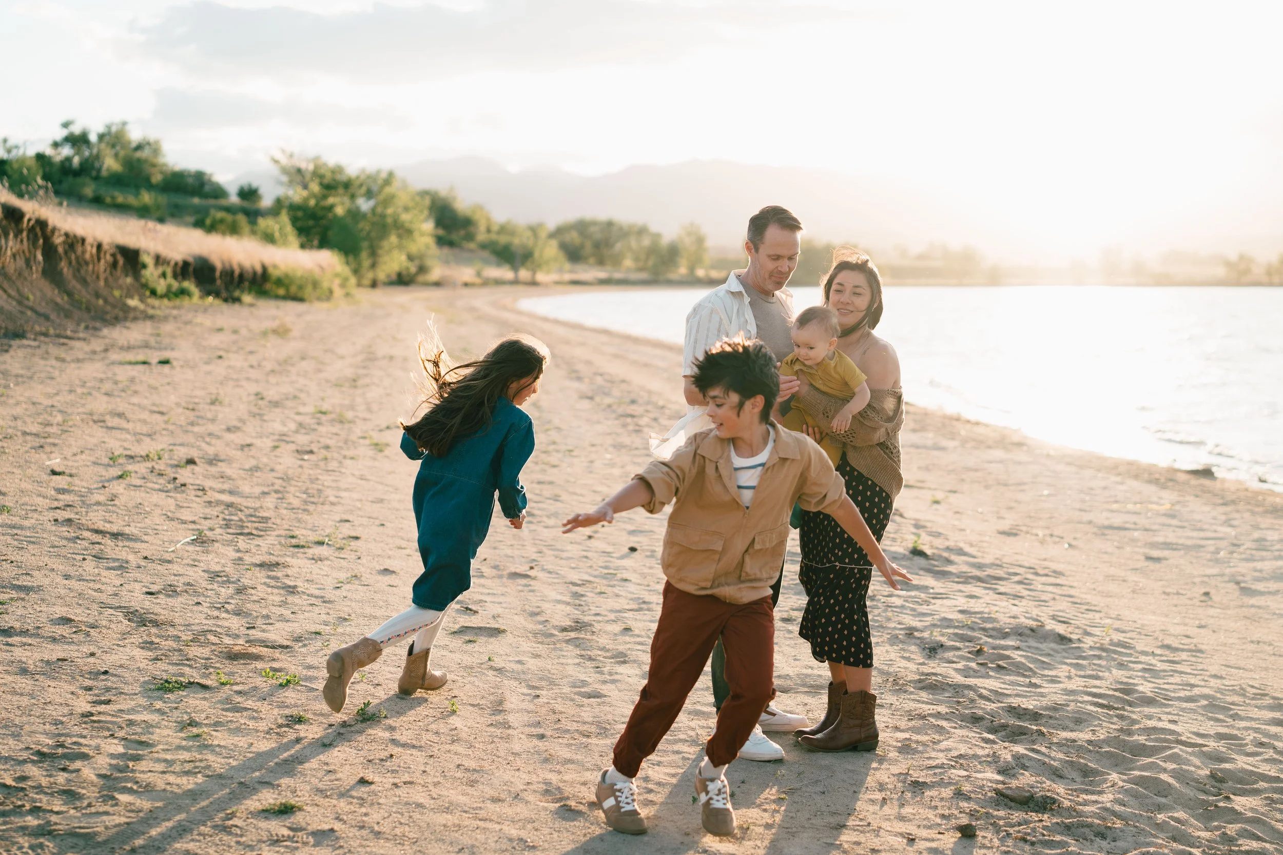 A group of children and adults playing together on a sandy beach near a body of water with trees in the background, during sunset or sunrise.