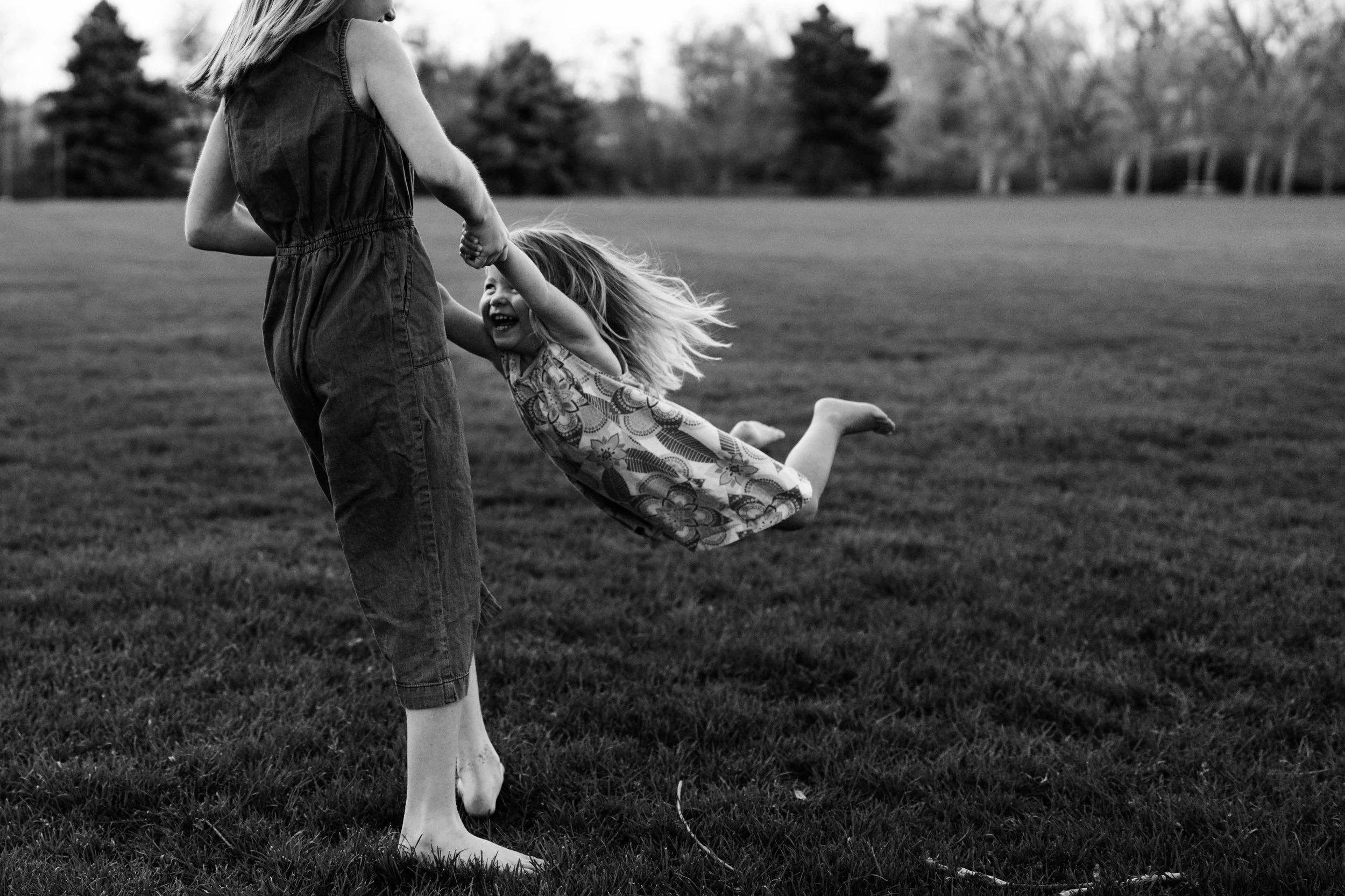 A black-and-white photo of a woman holding a young girl by the hands, spinning her in a grassy field with trees in the background. The girl is smiling and appears to be enjoying the outdoor activity.