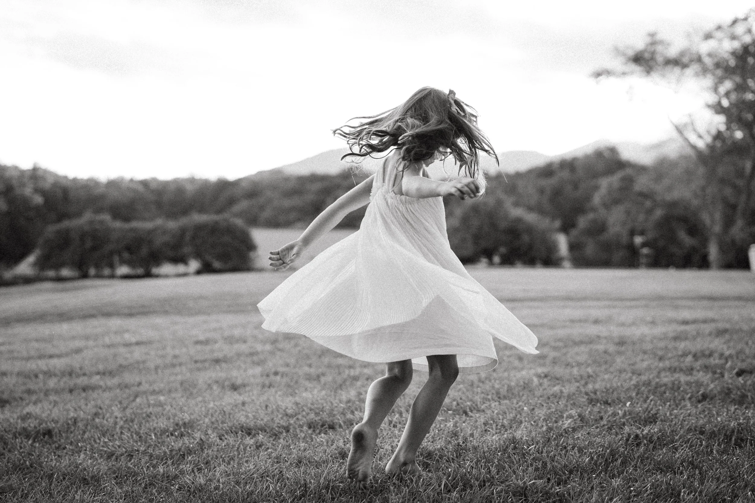 A girl in a light dress twirling and spinning on a grassy field outdoors, with mountain scenery and trees in the background, in black and white.