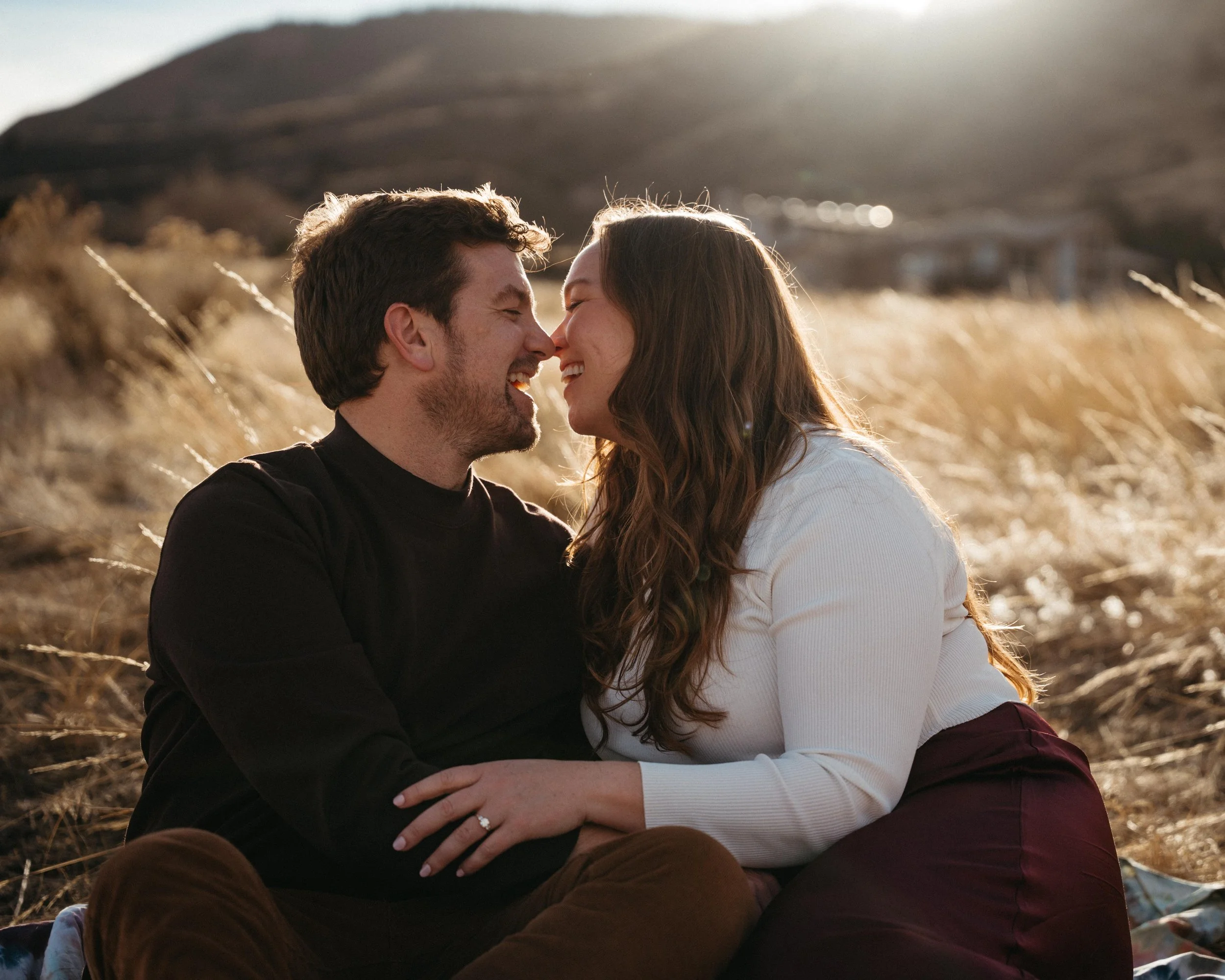 A couple with their foreheads touching and eyes closed, sitting outdoors in a field of tall grass with hills in the background, during what appears to be the late afternoon or sunset.