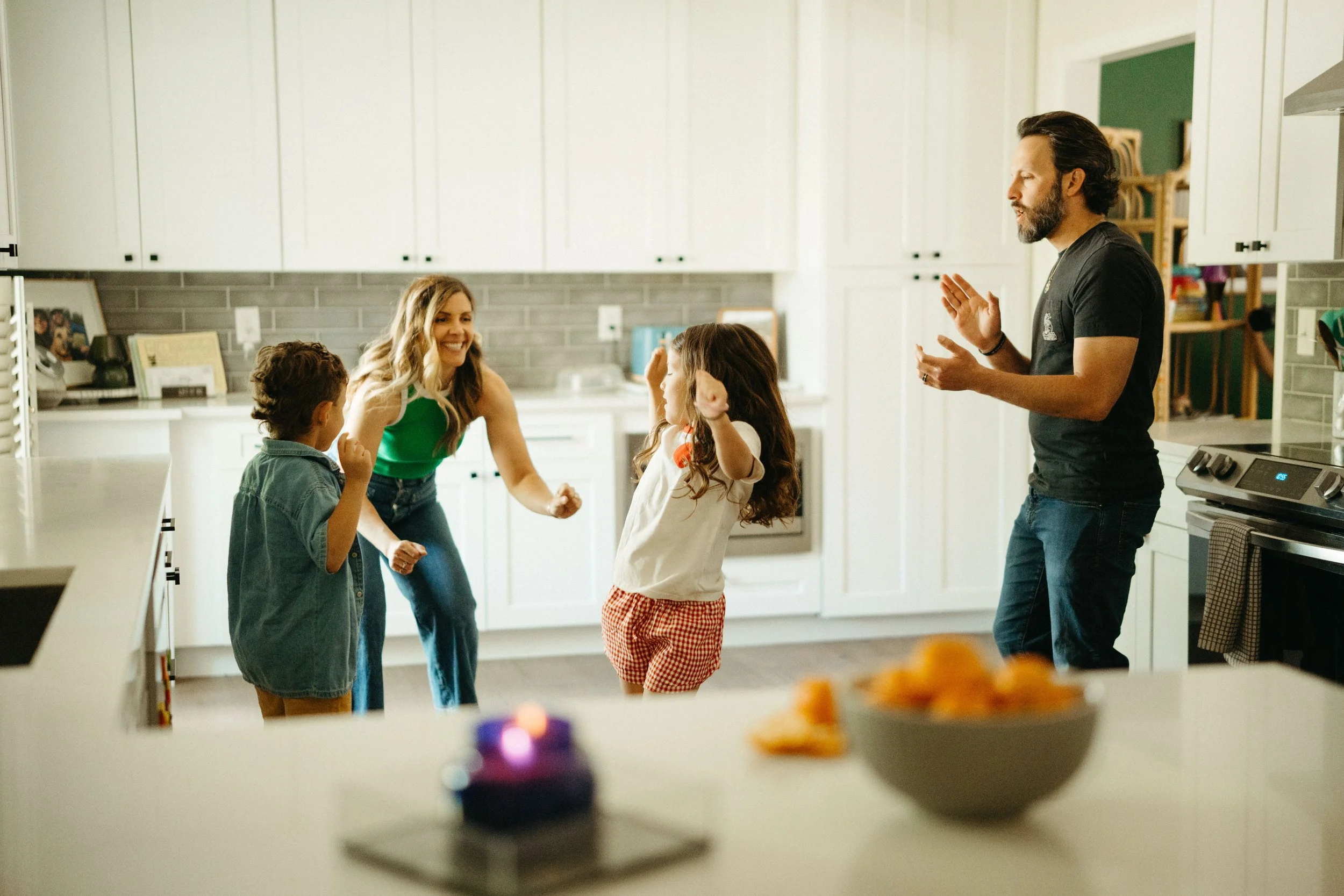 Family and friends dancing and celebrating in a kitchen with white cabinets and grey backsplash, with a birthday cake on the counter in the foreground.
